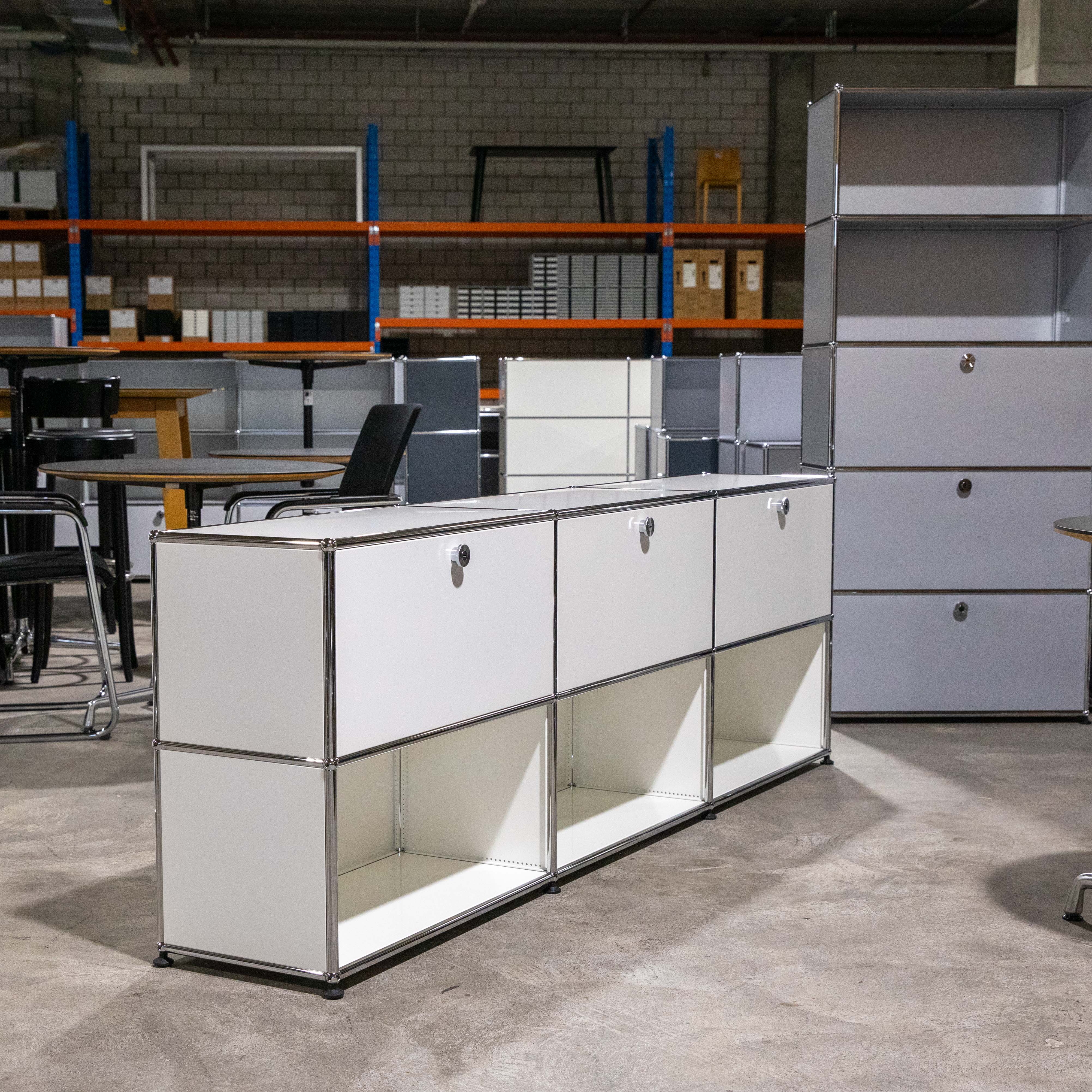 Weißes USM Haller Sideboard, modular: drei obere Fächer mit Klapptüren, unten drei offene Fächer. Glatte Oberfläche, typische Chromverbindungen. Steht in einem Lagerraum mit weiteren USM Möbeln und Regalen im Hintergrund.