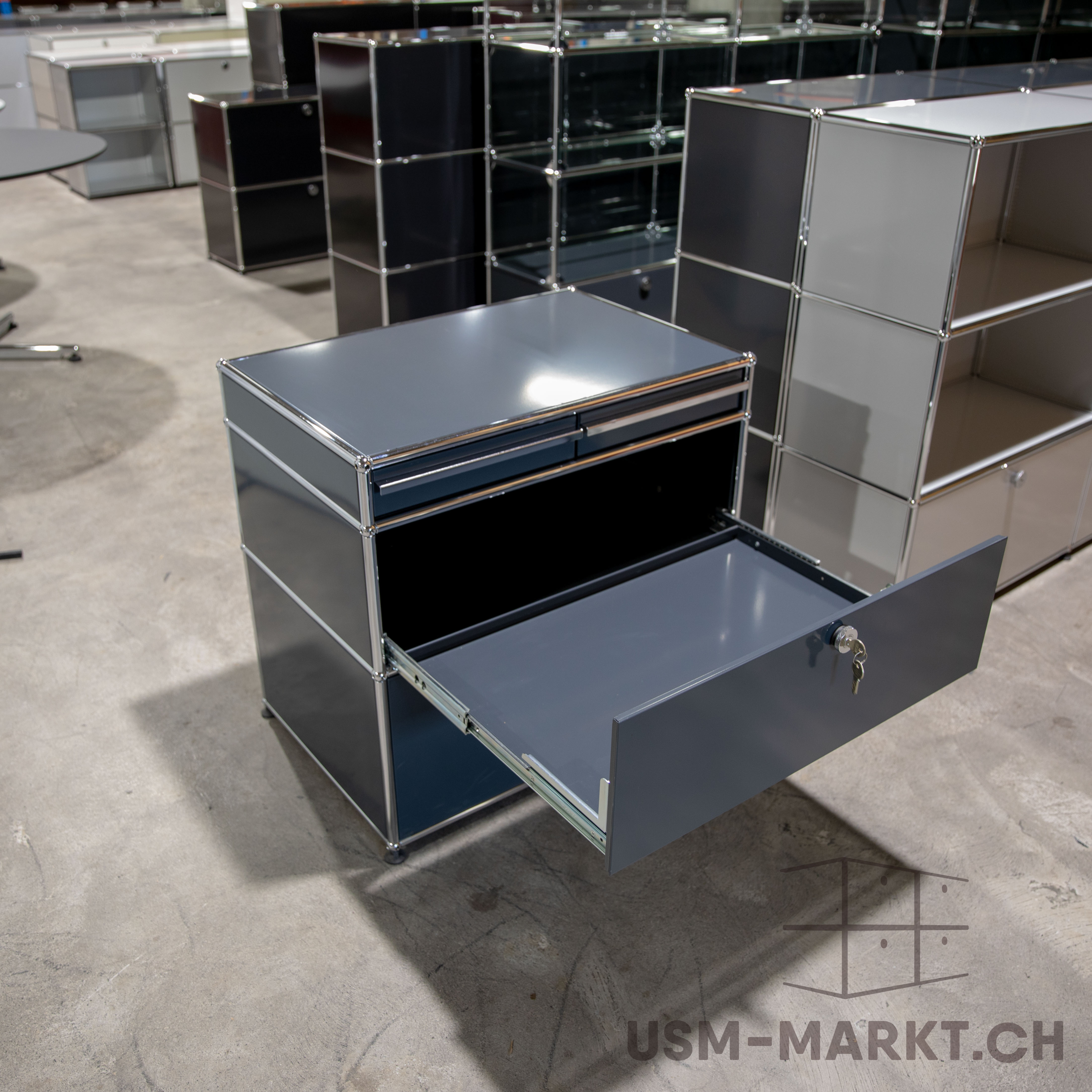 Dunkelgraues USM Haller Sideboard mit verchromten Verbindern auf Betonboden. Obere Schublade offen, ein Schlüssel hängt daran. Im Hintergrund weitere modulare Möbel in Grau, Schwarz und Weiß, wie in Lagerhalle oder Showroom.