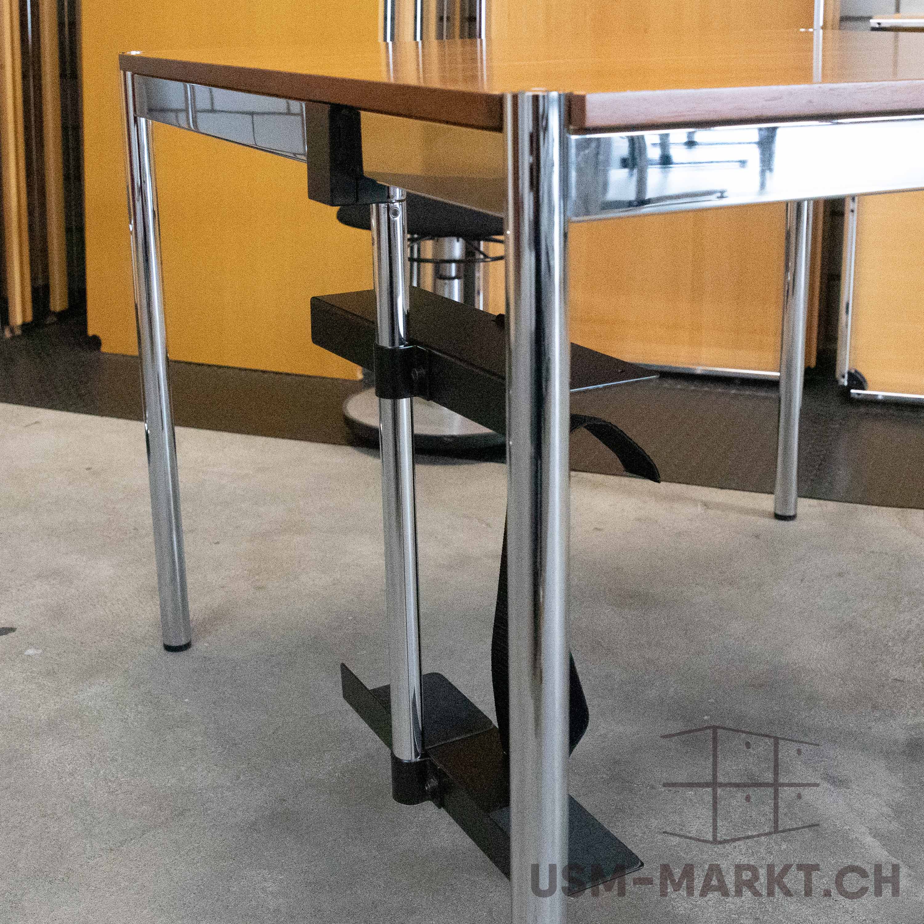 USM Haller table with brown wooden top and chrome-plated tubular steel legs on a light grey concrete floor. A black accessory is attached under the top, serving as cable management or a footrest. Yellow wall behind.