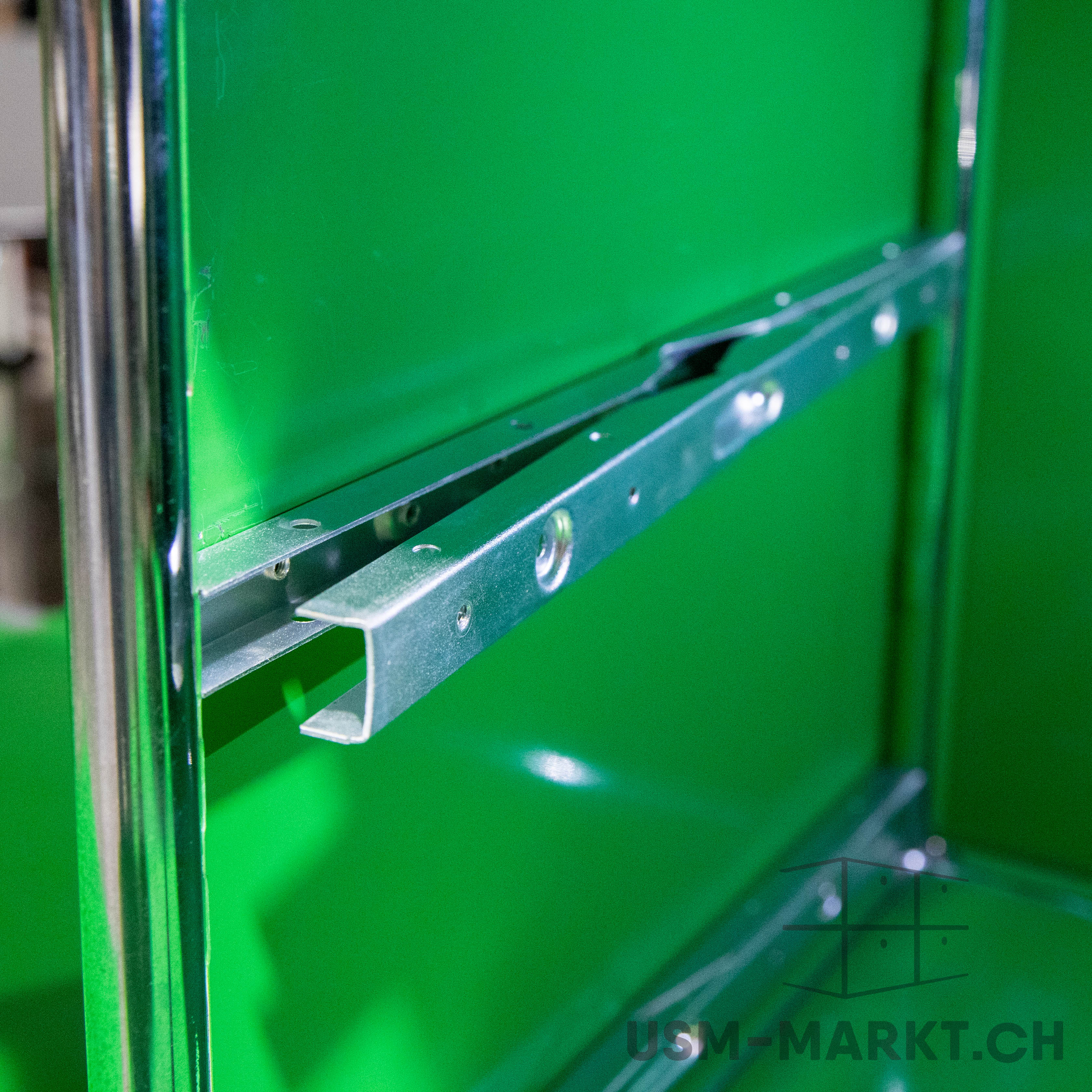 Interior view of a USM Haller unit in bright green. A metallic U-shaped guide rail is mounted horizontally on the back panel, ready for a drawer or shelf. Shiny chrome tubes on the left; textured green surface.