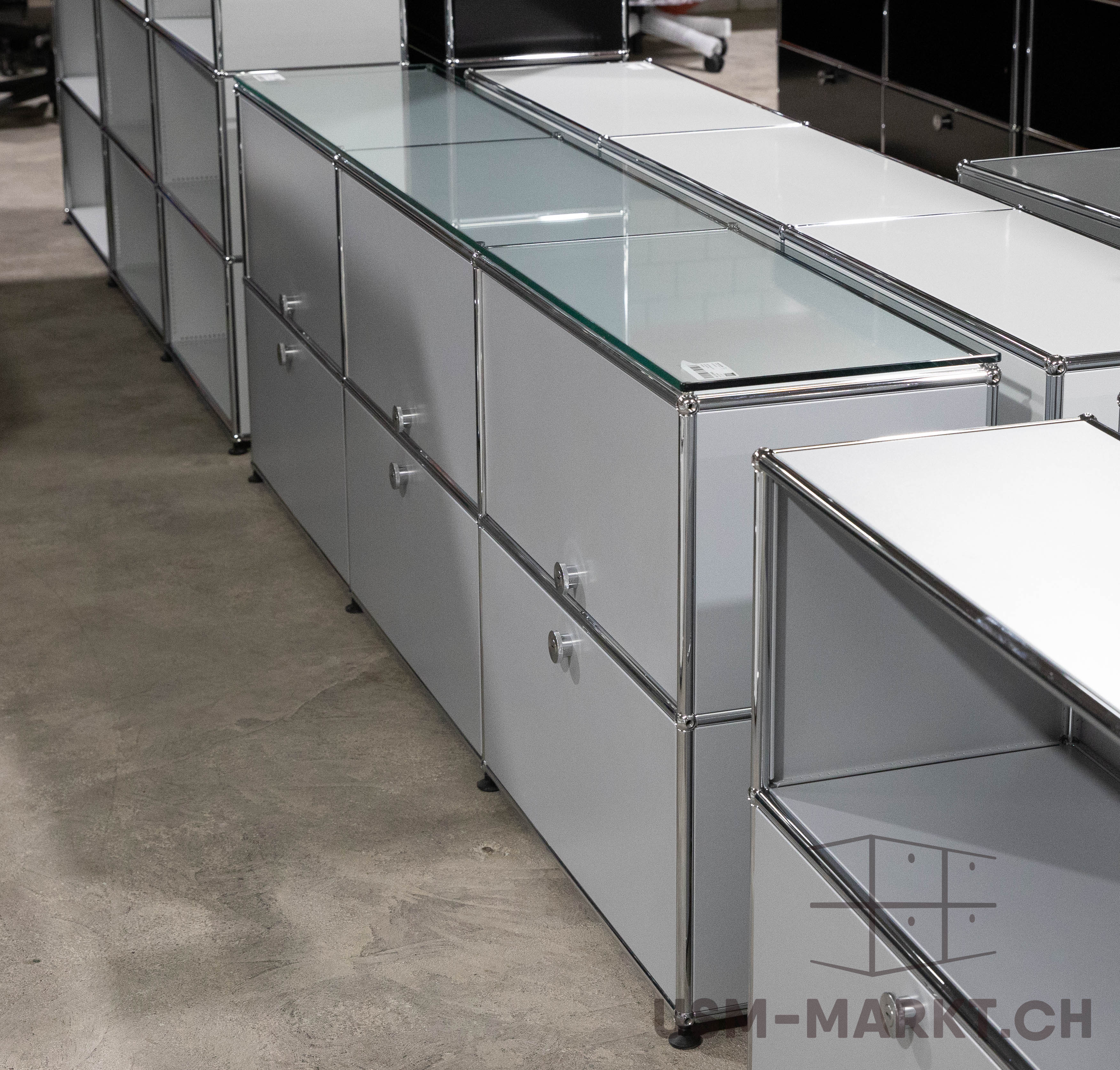 A set of modular light grey cupboards and shelves with polished chrome struts and fittings on a grey concrete floor. Some modules have drawers with round handles, whilst others feature transparent glass panels. The furniture has a modern, industrial look.