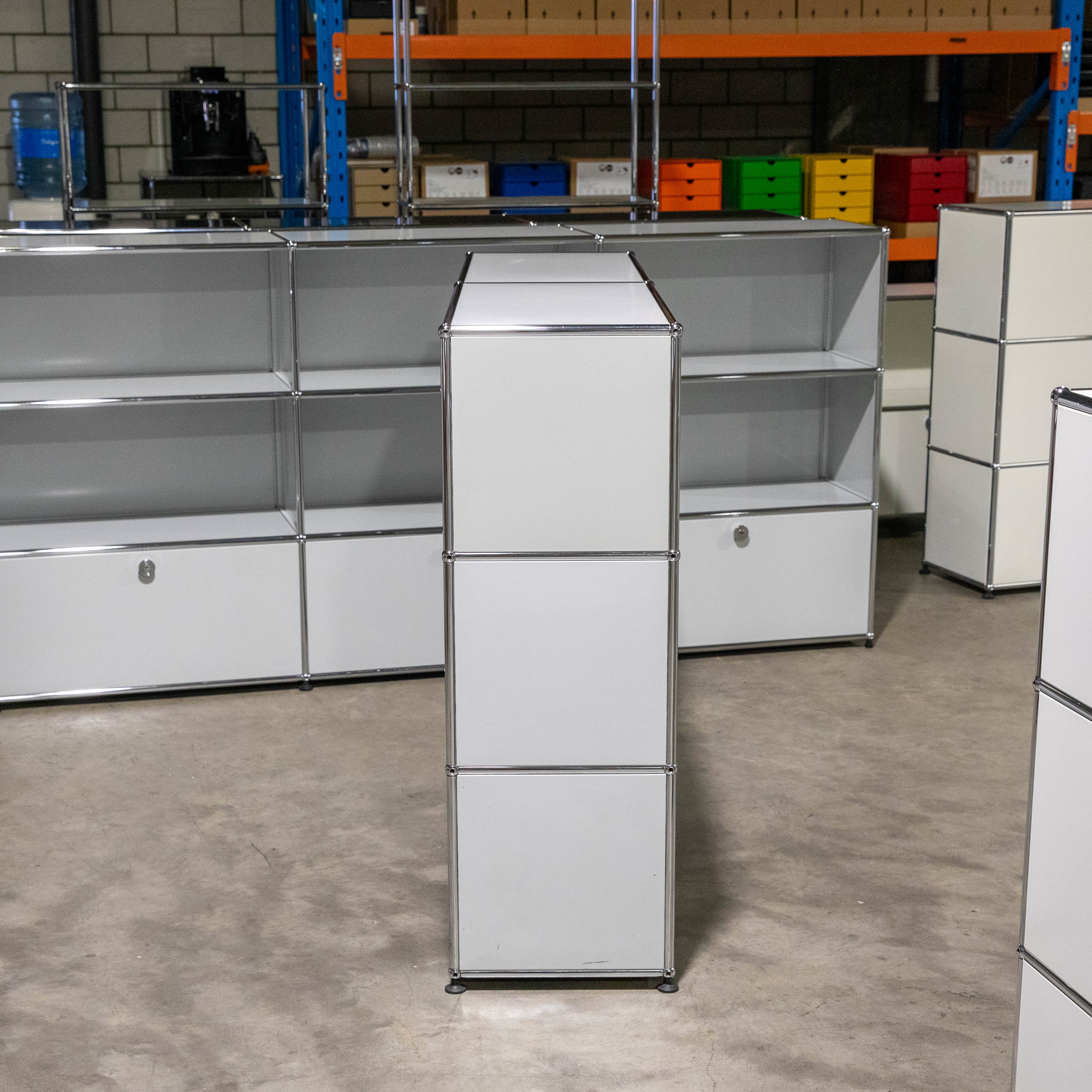 Wide, light grey USM Haller shelving with four open top compartments and two bottom-left closed drawers with handles. On a concrete warehouse floor. A narrow tall USM unit in front, with more shelving units behind.