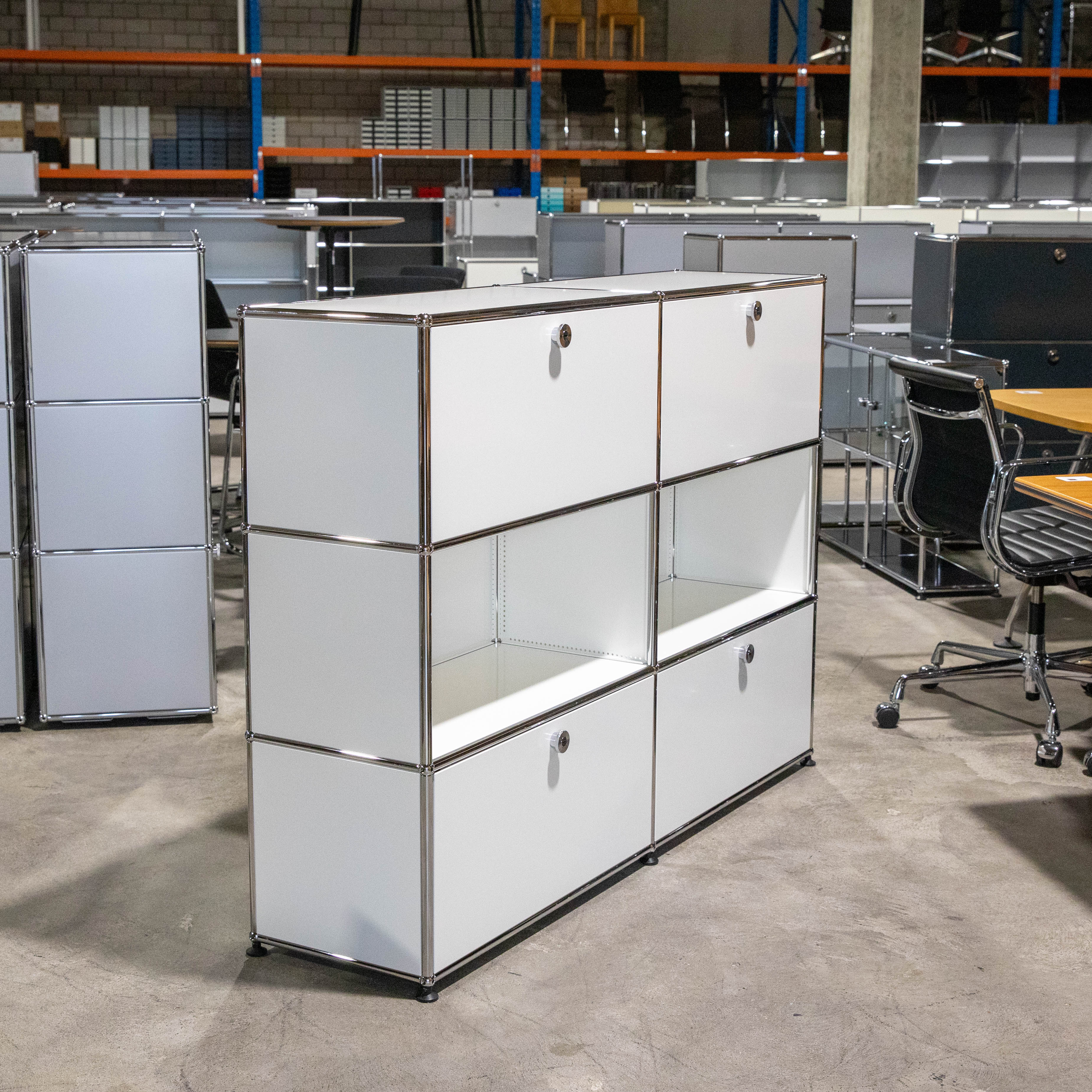Weisses USM Haller Regal, 2 Felder breit, 3 Fächer hoch, mit 4 geschlossenen Türen (Chromgriffe) und 2 offenen Fächern mittig. Modulare Struktur aus weissen Paneelen und Chromrohren auf grauem Betonboden. Hintergrund unscharf: Lagerregale, Büromöbel.
