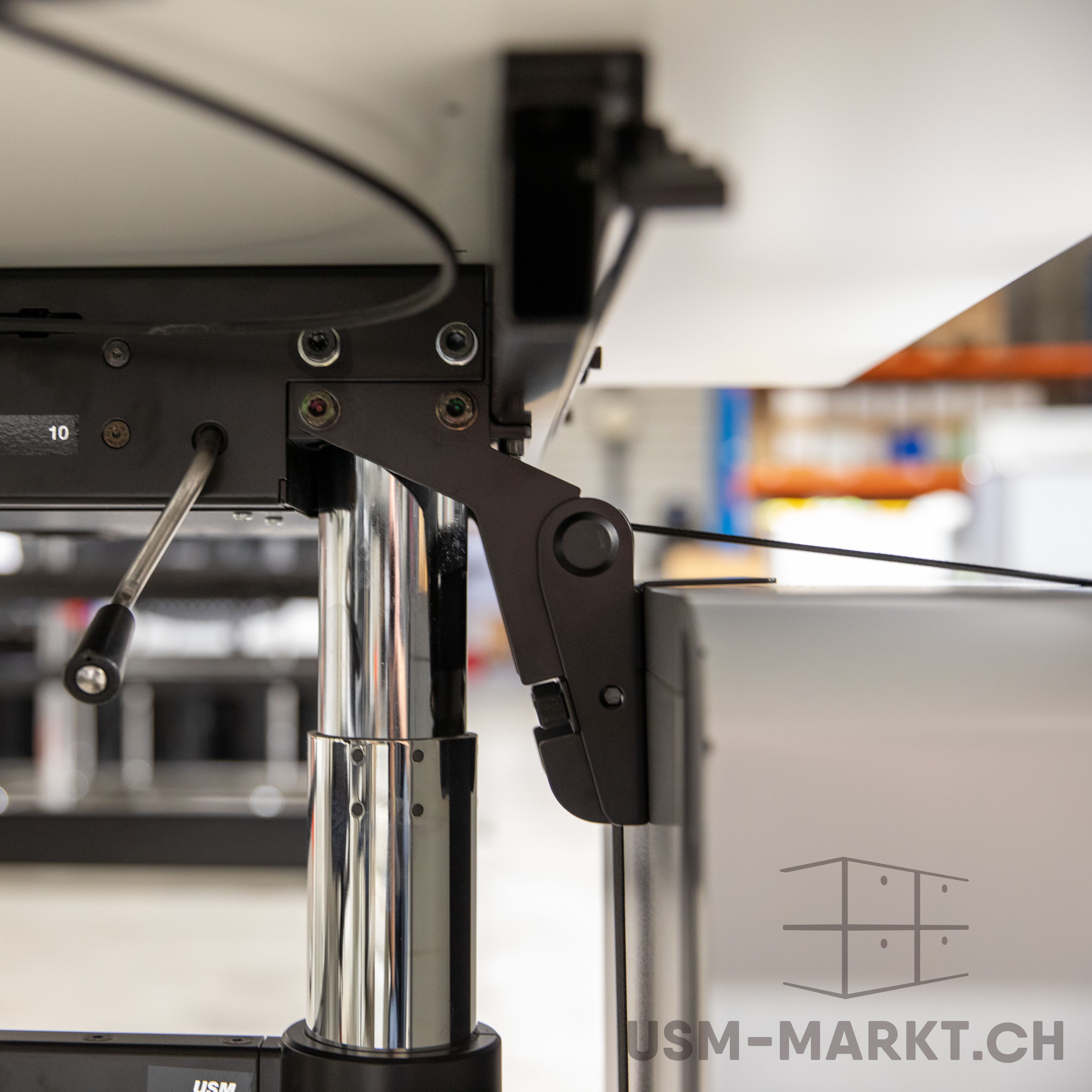 Nahaufnahme der Unterseite eines höhenverstellbaren Tisches: schwarzer Metallrahmen mit sichtbaren Schrauben und Zahl „10“. Verchromtes Tischbein und schwarzer Verstellhebel. Rechts schwarzer Gelenkmechanismus. Unscharfer Lagerhallenhintergrund.