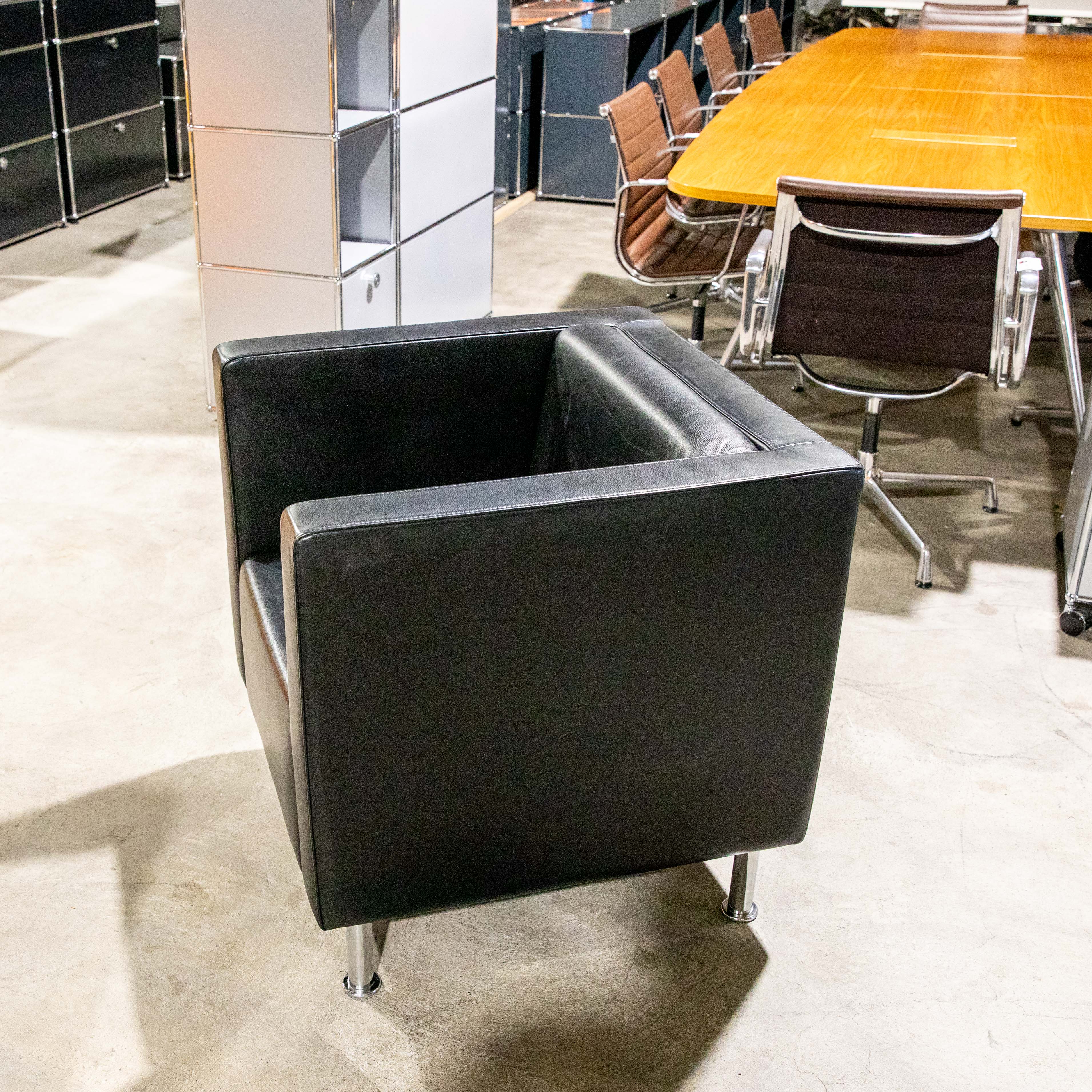A black leather armchair on a light-coloured concrete floor in the foreground. Behind it, a long, polished wooden table with several brown, upholstered office chairs with metal frames. Modular black and white cabinets at the back on the left.