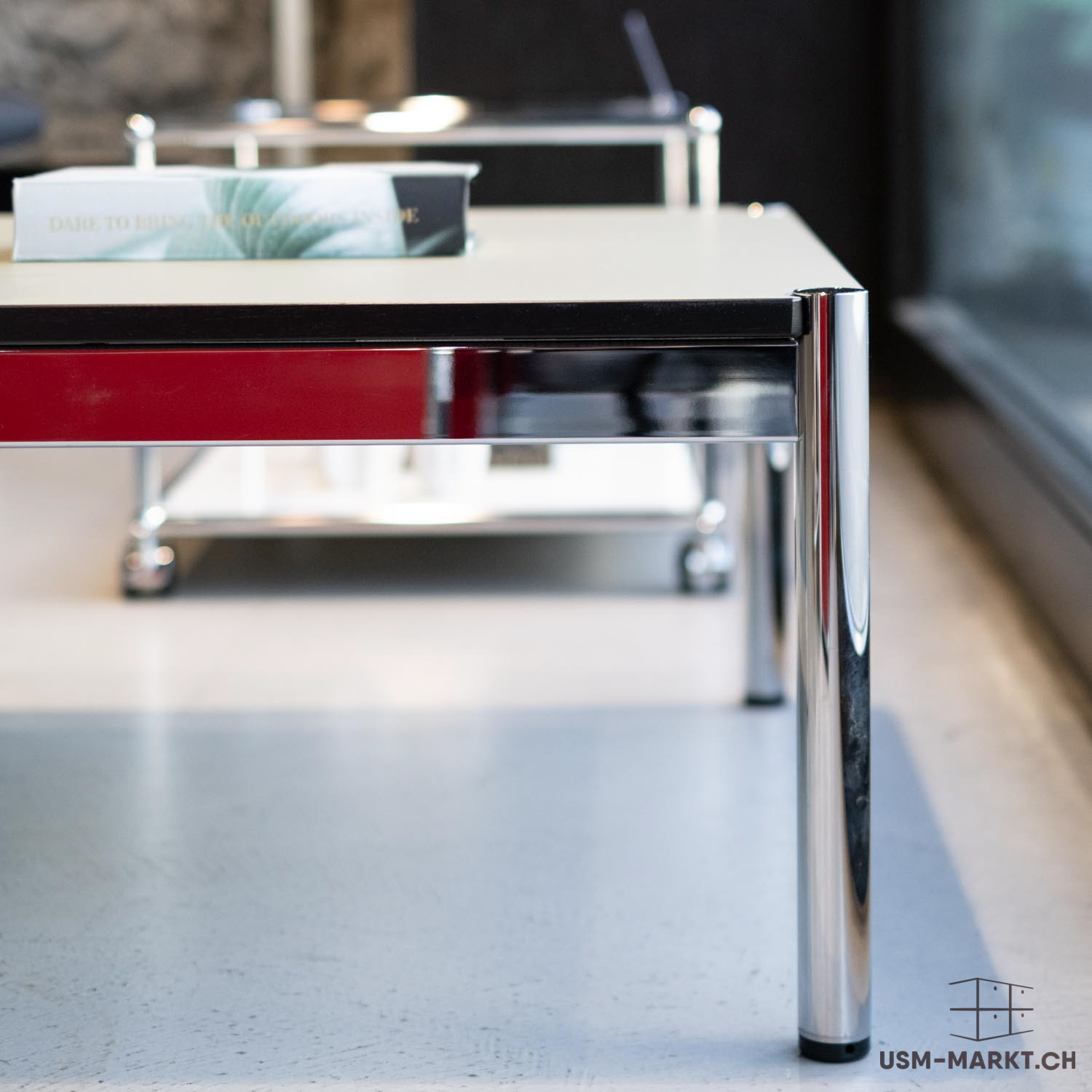 Mobile USM Haller con piano del tavolo chiaro, frontale rosso brillante e gambe cromate. Sul piano è appoggiato un libro con un motivo di foglie verdi e la scritta "DARE TO BRING THE OUTDOORS INSIDE". Tubi cromati a vista, su uno sfondo chiaro.