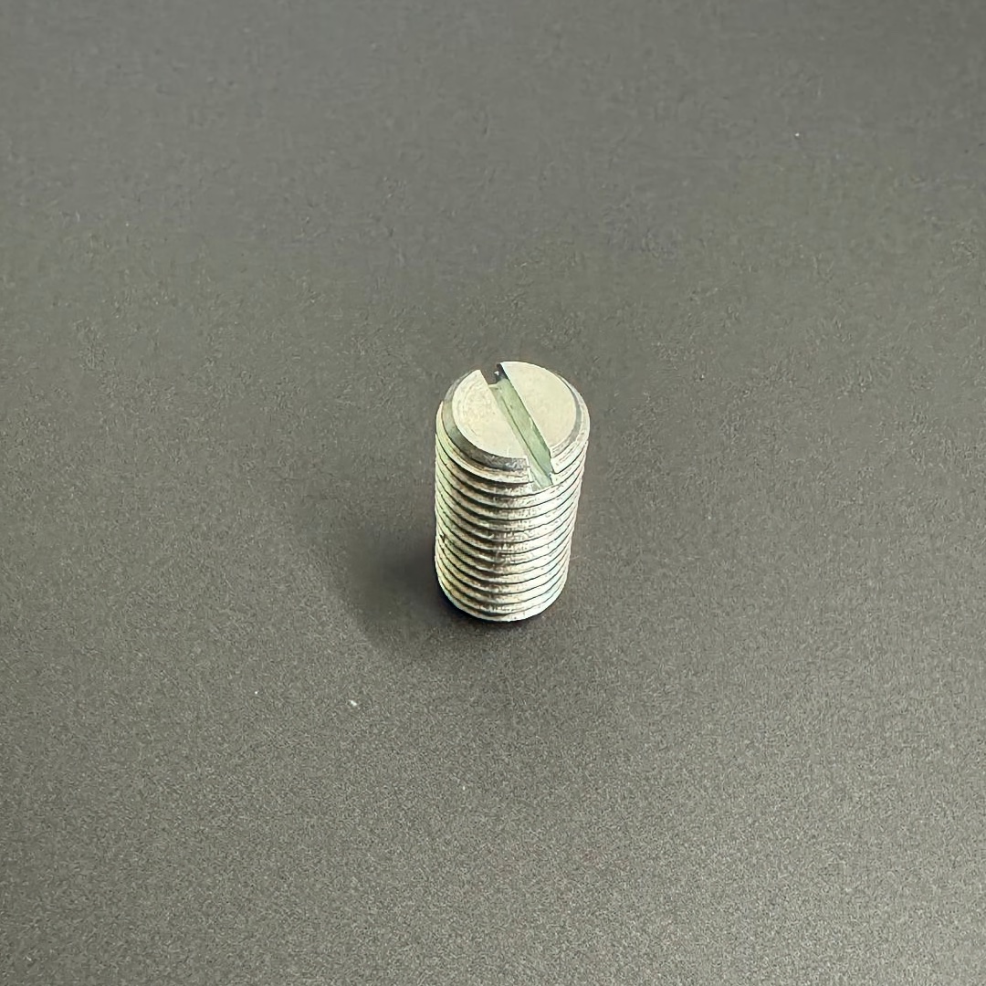 Ein einzelner, zylindrischer Gewindestift aus Metall steht aufrecht auf einer dunklen Oberfläche. Er hat ein durchgehendes Außengewinde und oben einen Schlitz für einen Flachschraubendreher. Die Metalloberfläche ist glänzend und silbern.