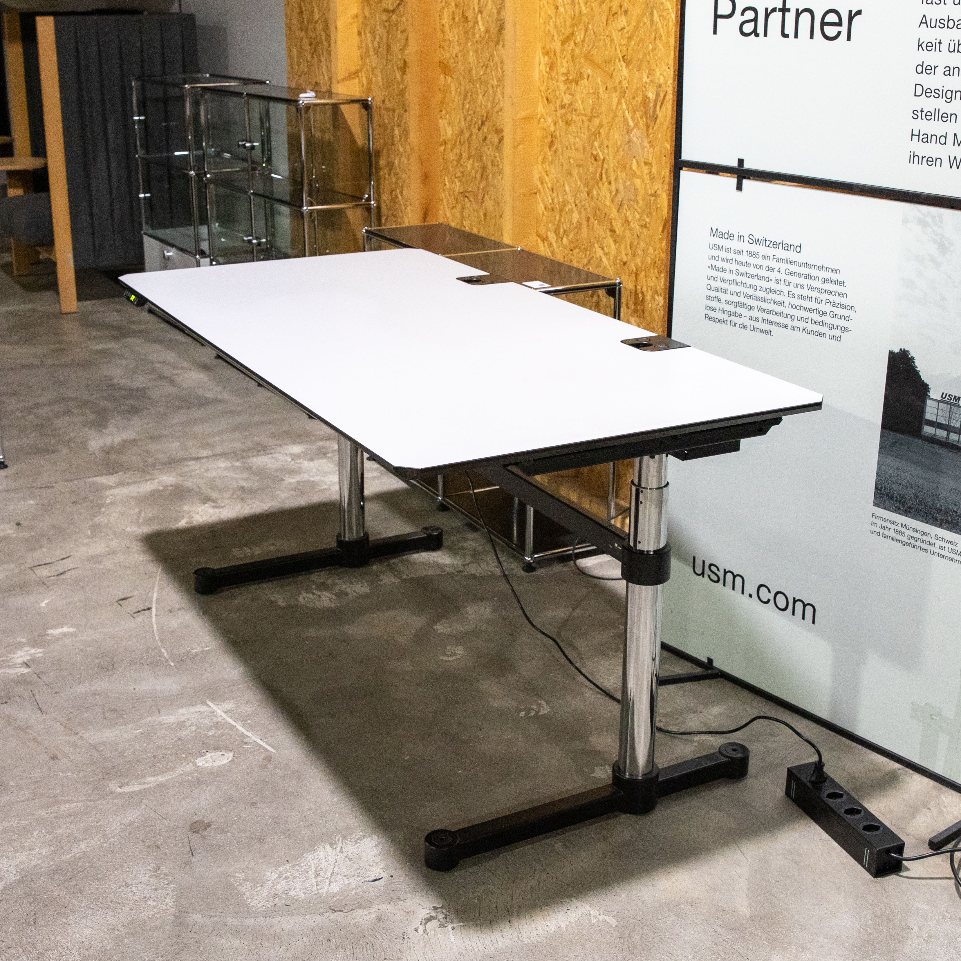Elektrisch höhenverstellbarer USM Kitos E Plus Schreibtisch mit weisser Kunstharzplatte und abgerundeten Ecken. Auf zwei verchromten Beinen mit schwarzen Fußgestellen, mit zwei runden Kabeldurchführungen. Hintergrund: Holzspanplatte, Regal.