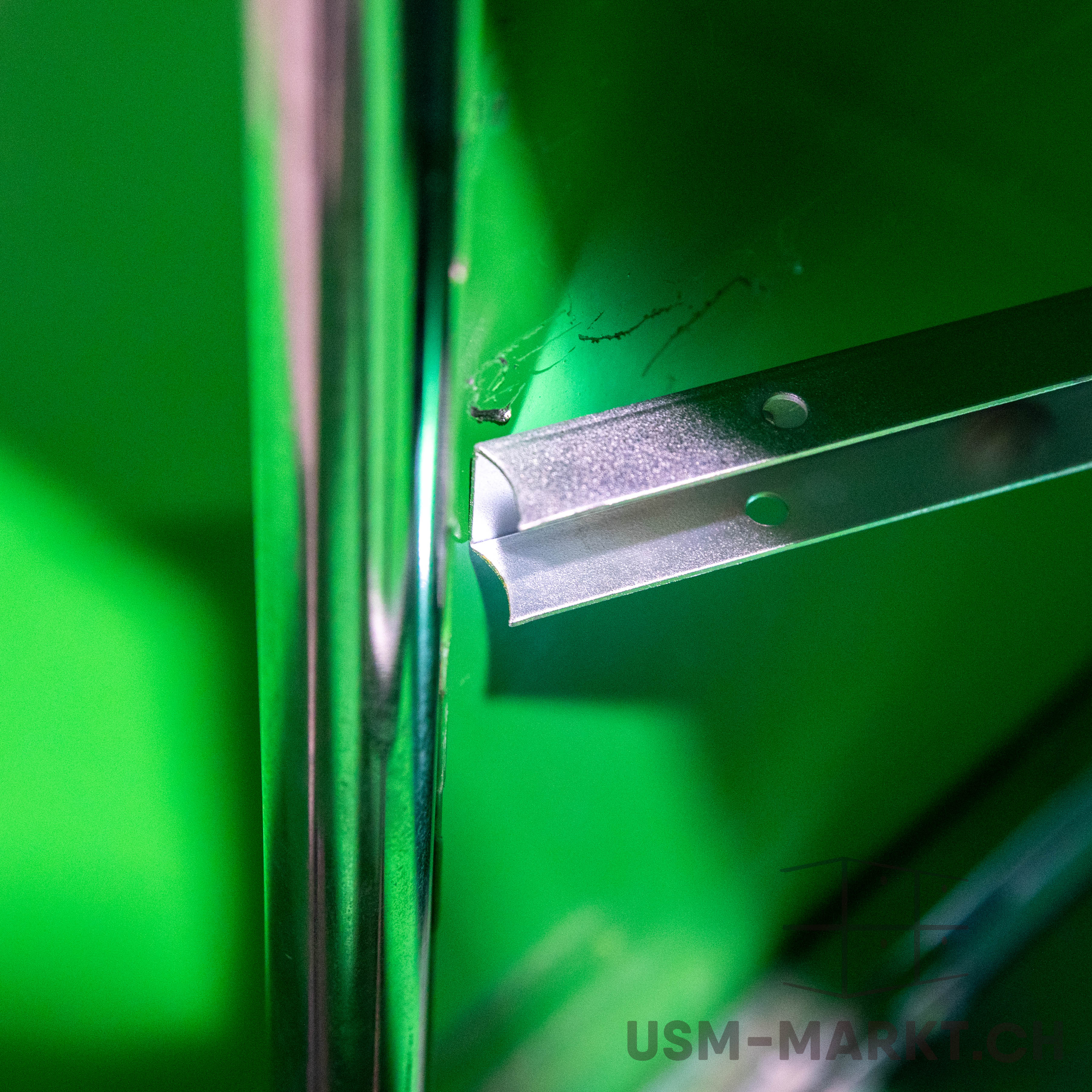 Close-up of a shiny chrome rod on the left, set against a bright green background. A silver U-shaped metal profile is attached horizontally to the green wall, with two holes. Slight dark marks are visible.