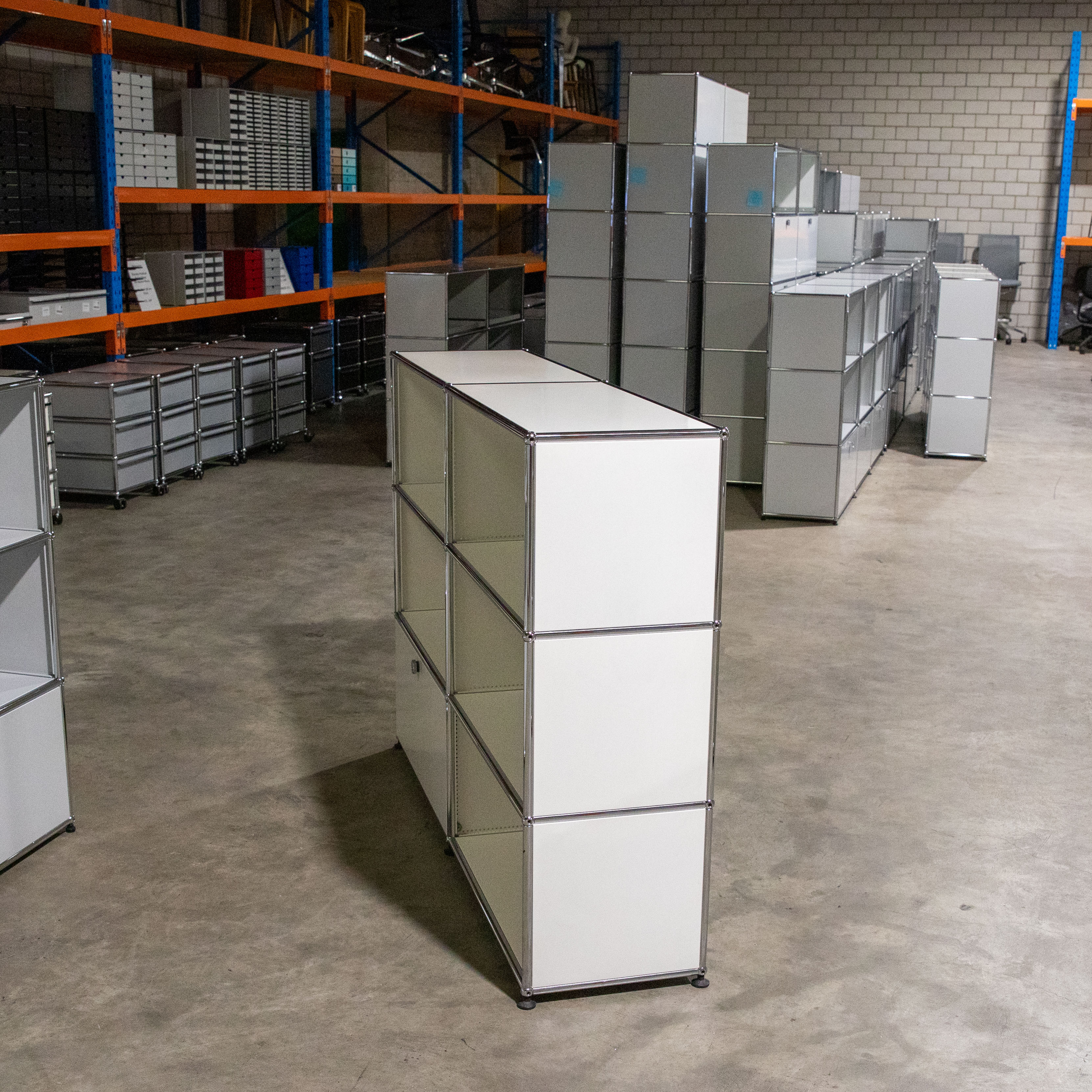 Weißes, gebrauchtes USM Haller Regal, 2 Felder breit, 3 Fächer hoch, teils offen, teils geschlossen. Modulares Möbel mit verchromten Stahlrohren und weißen Metallplatten auf Betonboden. Hintergrund: weitere USM Möbel, orange Lagerregale.