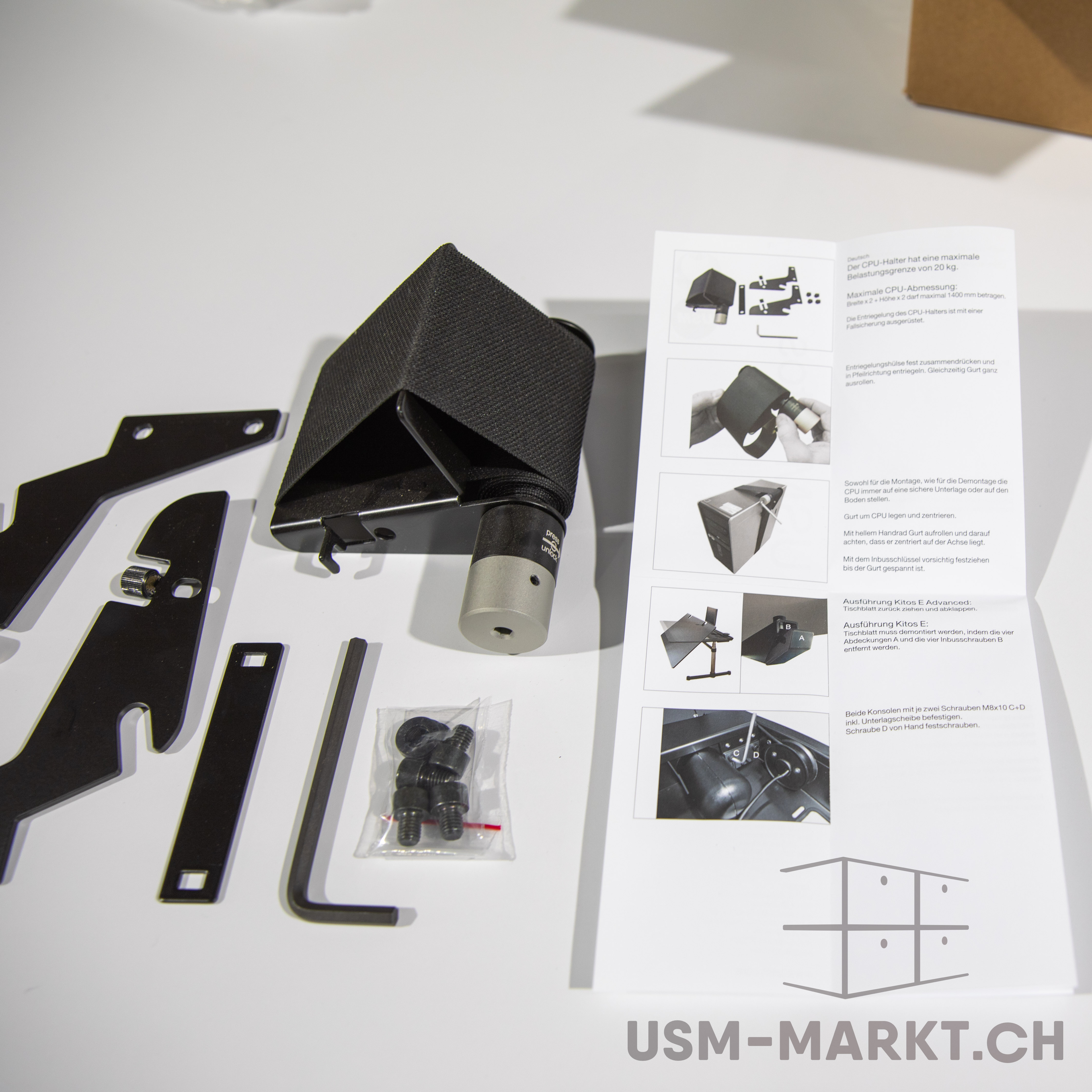 Ein flaches Arrangement zeigt die schwarzen Einzelteile eines CPU-Halters: Metallwinkel, Schrauben und einen Inbusschlüssel. Rechts daneben liegt eine Anleitung mit Bildern und deutschem Text zur Montage und Handhabung.