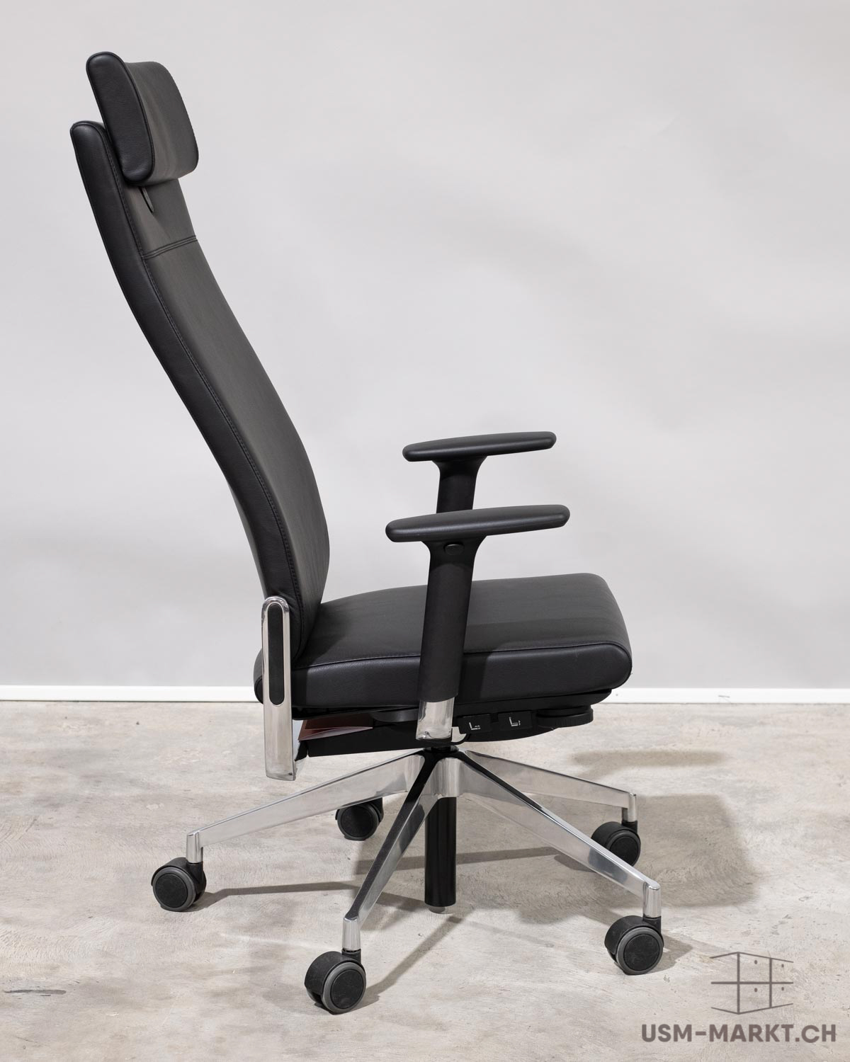 Fauteuil de bureau noir à dossier haut, avec appui-tête intégré et accoudoirs réglables, vu de profil. Piètement à cinq branches argenté brillant avec roulettes noires. Assise et dossier ergonomique en cuir noir, posé sur un sol en béton gris clair.