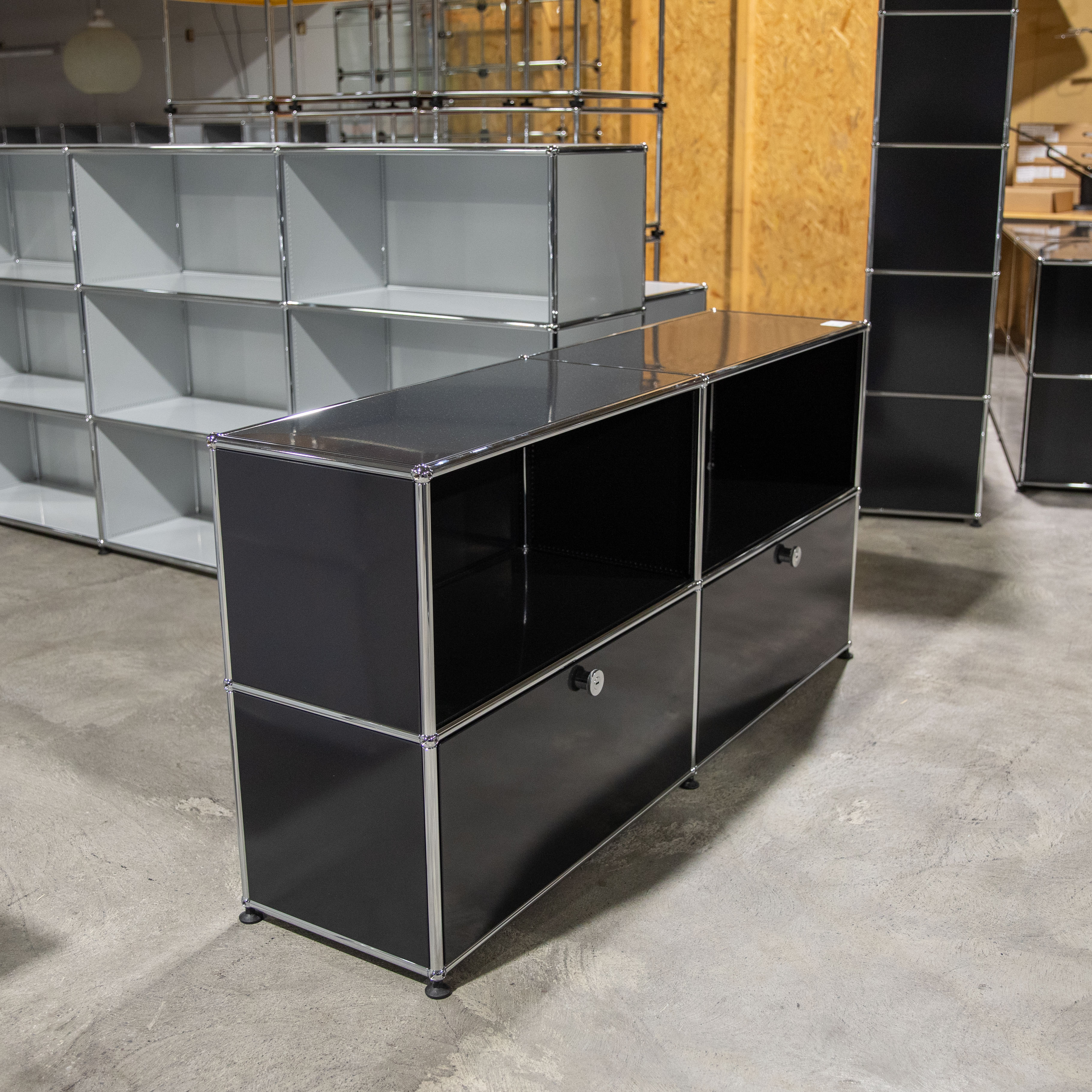 Ein schwarzes, modulares Sideboard mit glänzenden Chromrahmen steht auf hellgrauem Betonboden. Es hat zwei offene Fächer oben und zwei Schubladen mit silbernen Griffen unten. Im Hintergrund sind weitere Regalmodule und eine Holzwand.