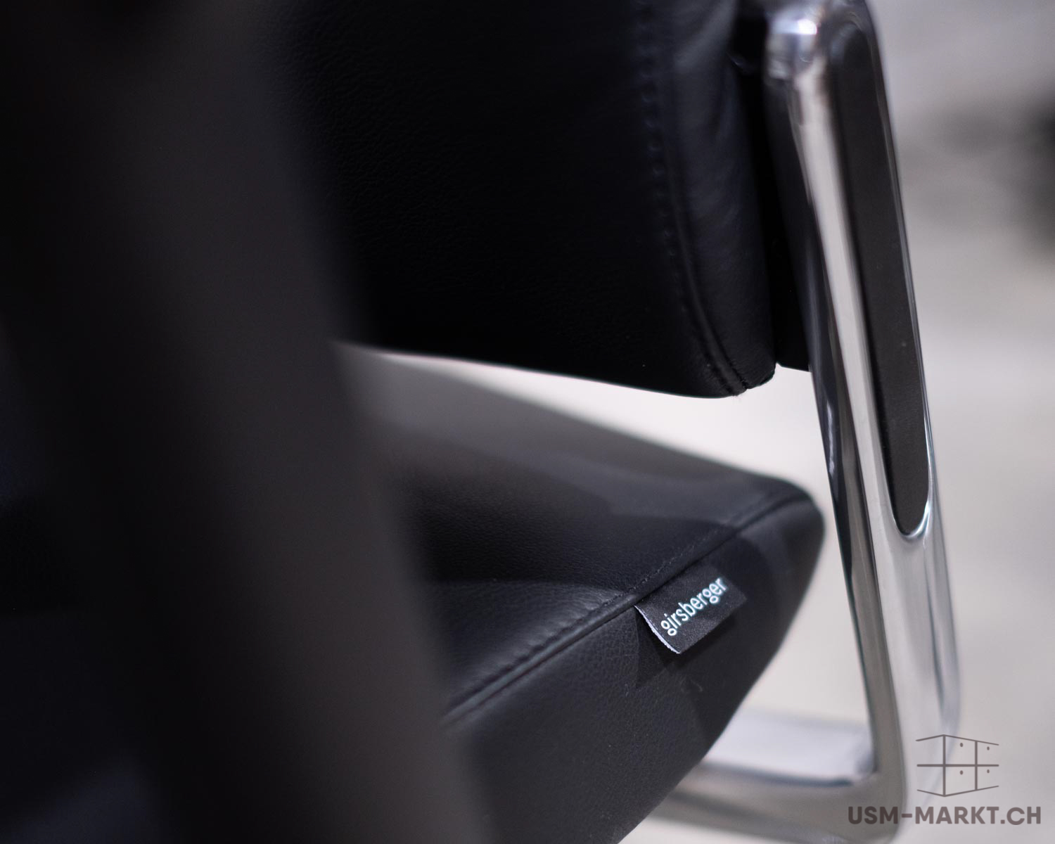 Vue détaillée d'un élégant siège noir « Girsberger » avec revêtement en cuir et structure chromée. Le nom de la marque « Girsberger » est visible sur une petite étiquette noire située à l'avant de l'assise, dans le cuir grainé.