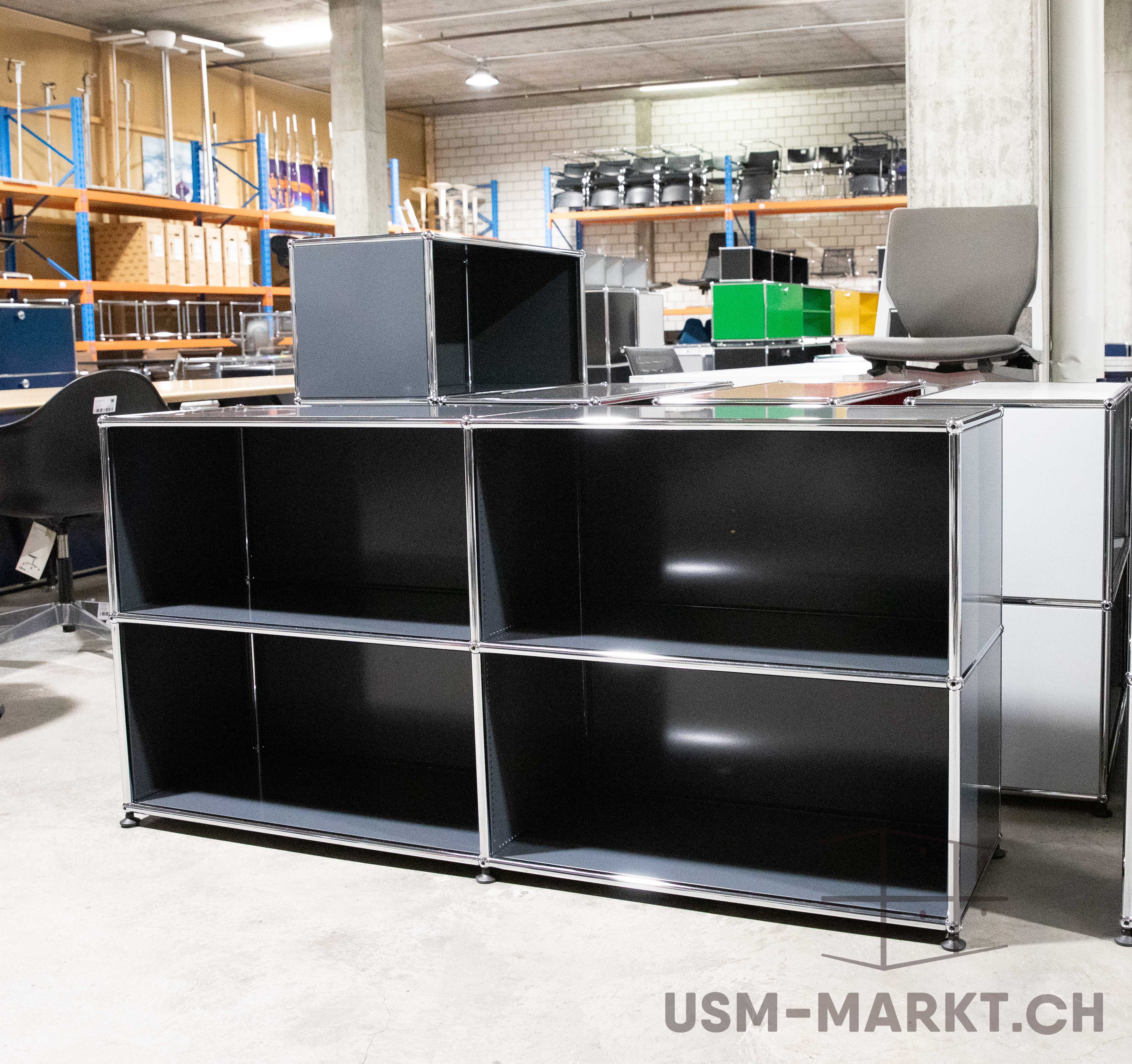 Schwarzes, modulares USM Haller Sideboard mit sechs offenen Fächern (2 Reihen à 3) auf hellem Betonboden. Glänzende Chromverbindungen und schwarze Stellfüße. Im Hintergrund Lagerregale mit Büromöbeln und Stühlen.
