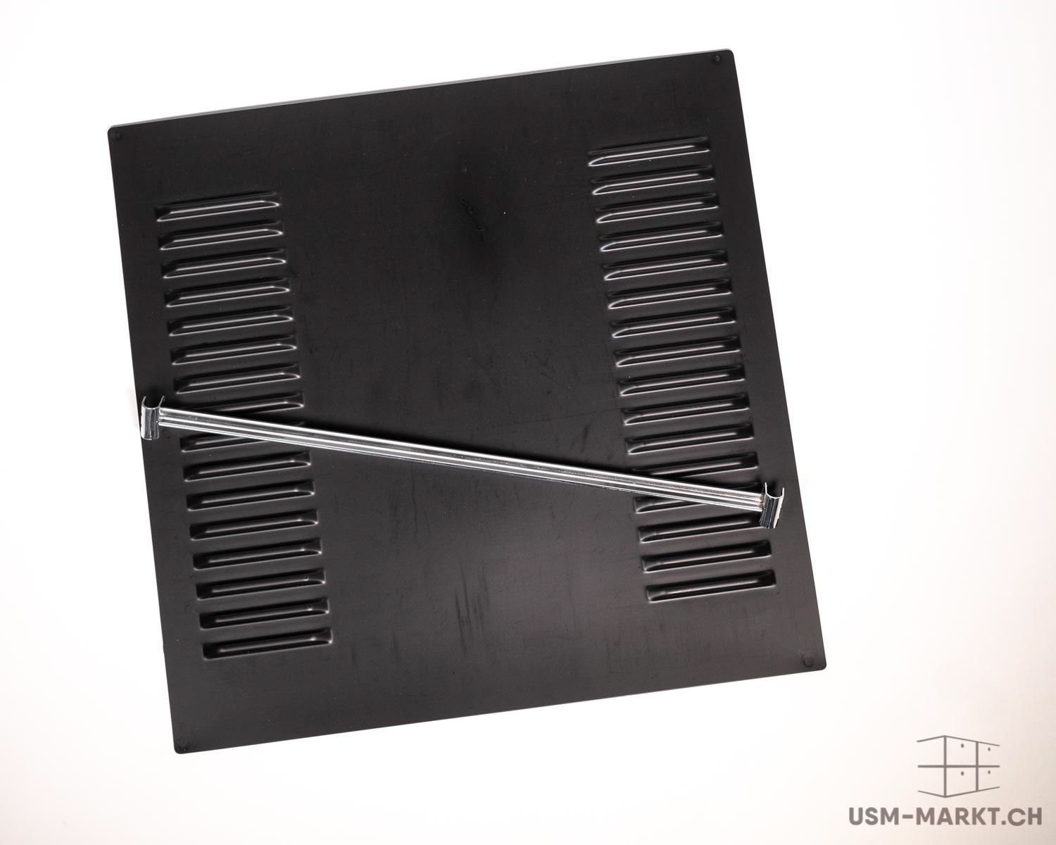 A black, rectangular ventilation panel with two vertical rows of horizontal slats. Across the top, a shiny, silver metal bar, fixed at both ends and positioned slightly diagonally, serving as a handle or support.