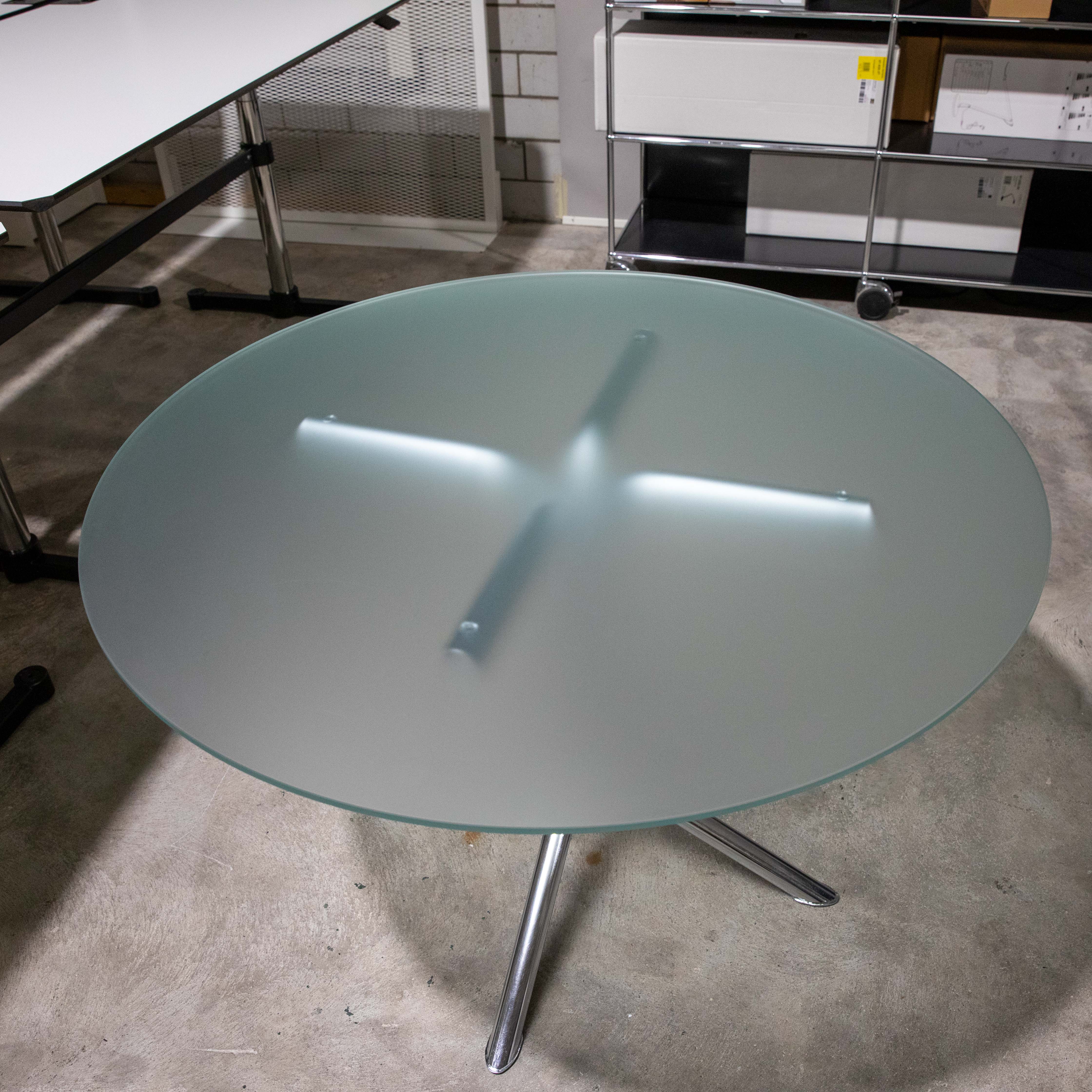 Table ronde avec plateau en verre dépoli et piètement à trois pieds chromé brillant. Le plateau est légèrement éclairé de l'intérieur et laisse apparaître la fixation en forme de croix. Sur un sol en béton gris, avec des meubles en arrière-plan.