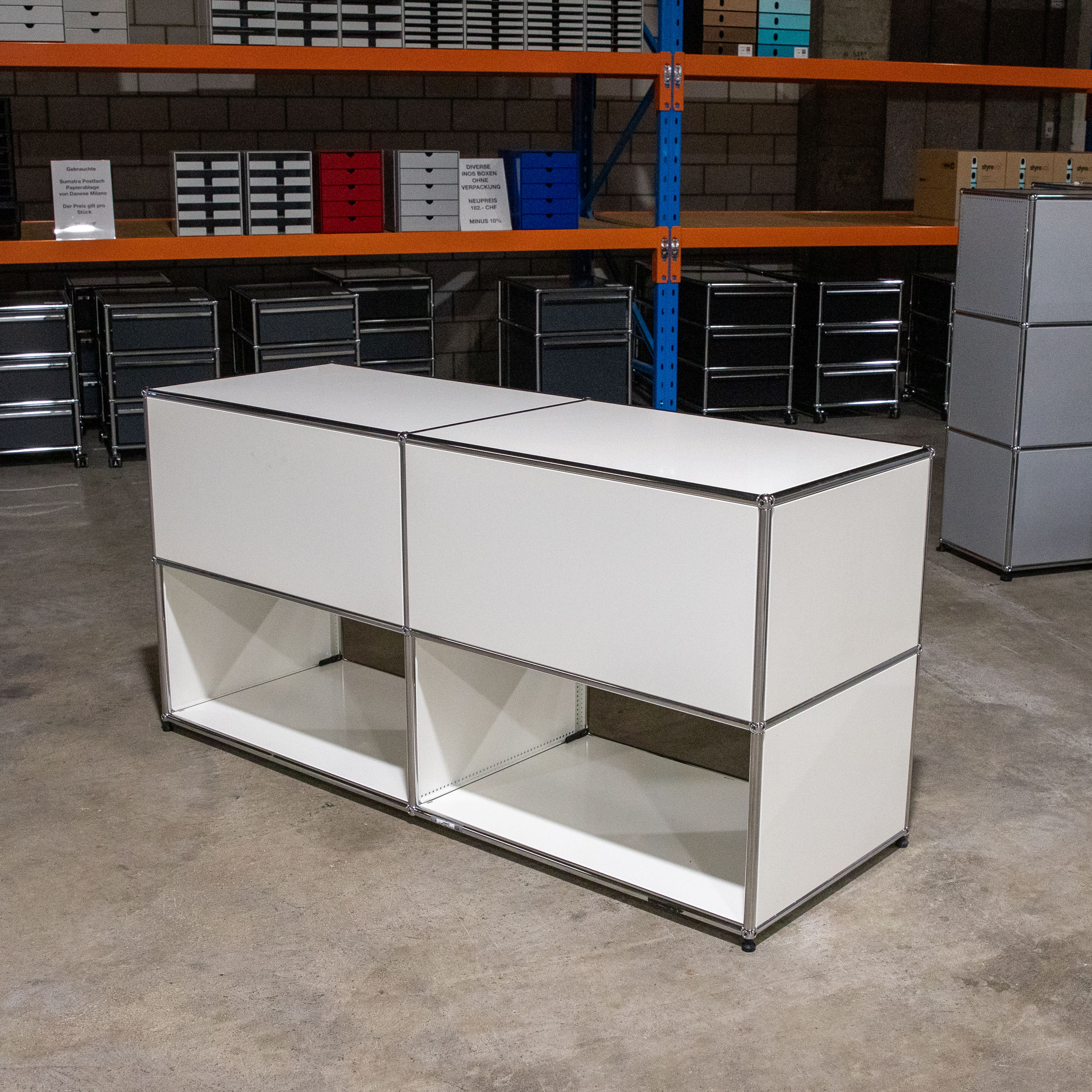 Weißes USM Haller Sideboard 2x2 mit verchromtem Stahlrohrrahmen. Obere 2 Fächer geschlossen, untere 2 offen, da die Einschubtürfronten fehlen. Steht auf Betonboden im Lagerraum, weitere USM Möbel hinten.