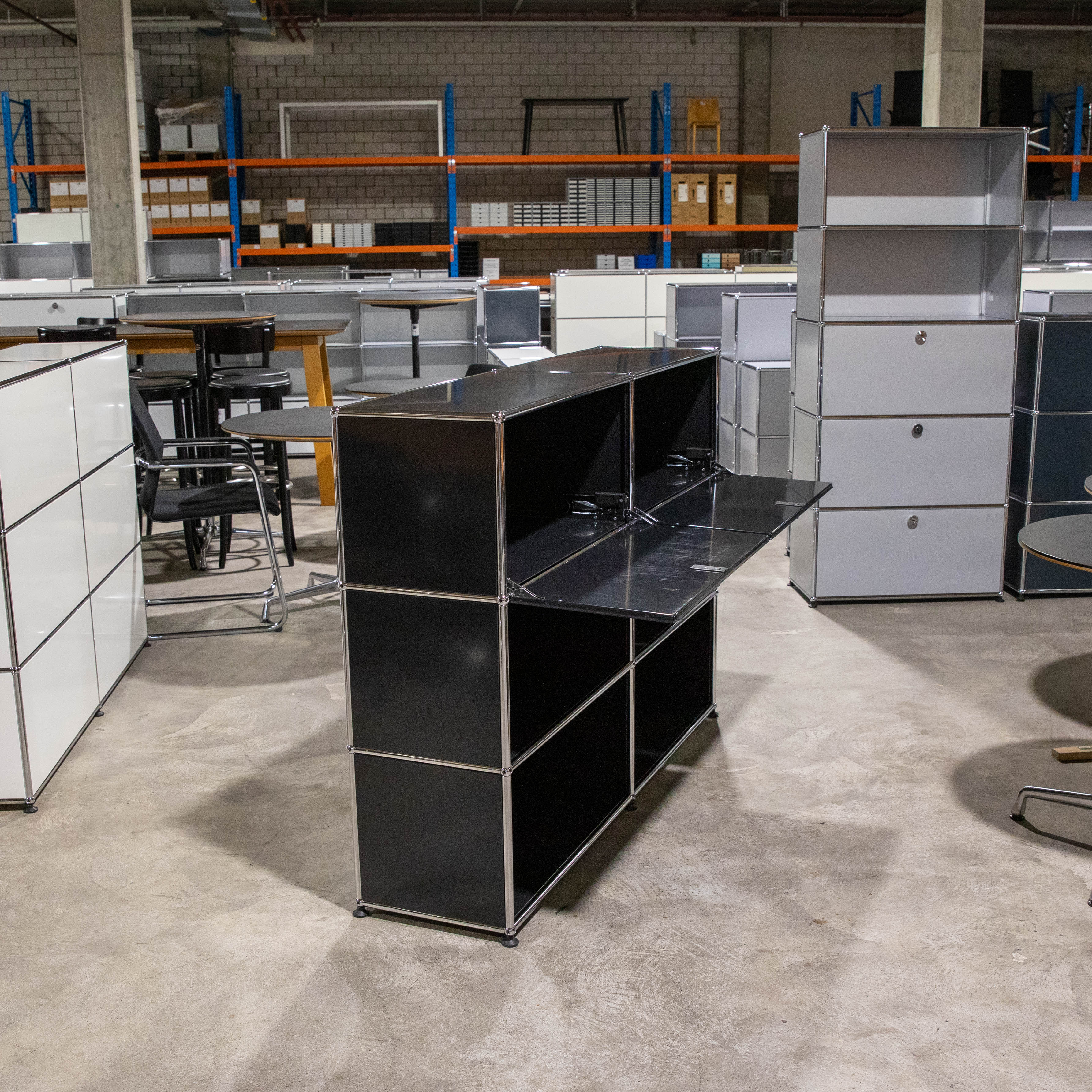 Ein elegantes USM Haller Regal in Schwarz steht zentral auf Betonboden, modulare 2x3-Konfiguration mit zwei oberen, leicht geöffneten Klapptüren, die Innenraum und Scharniere zeigen. Im Hintergrund USM Möbel und Lagerregale.