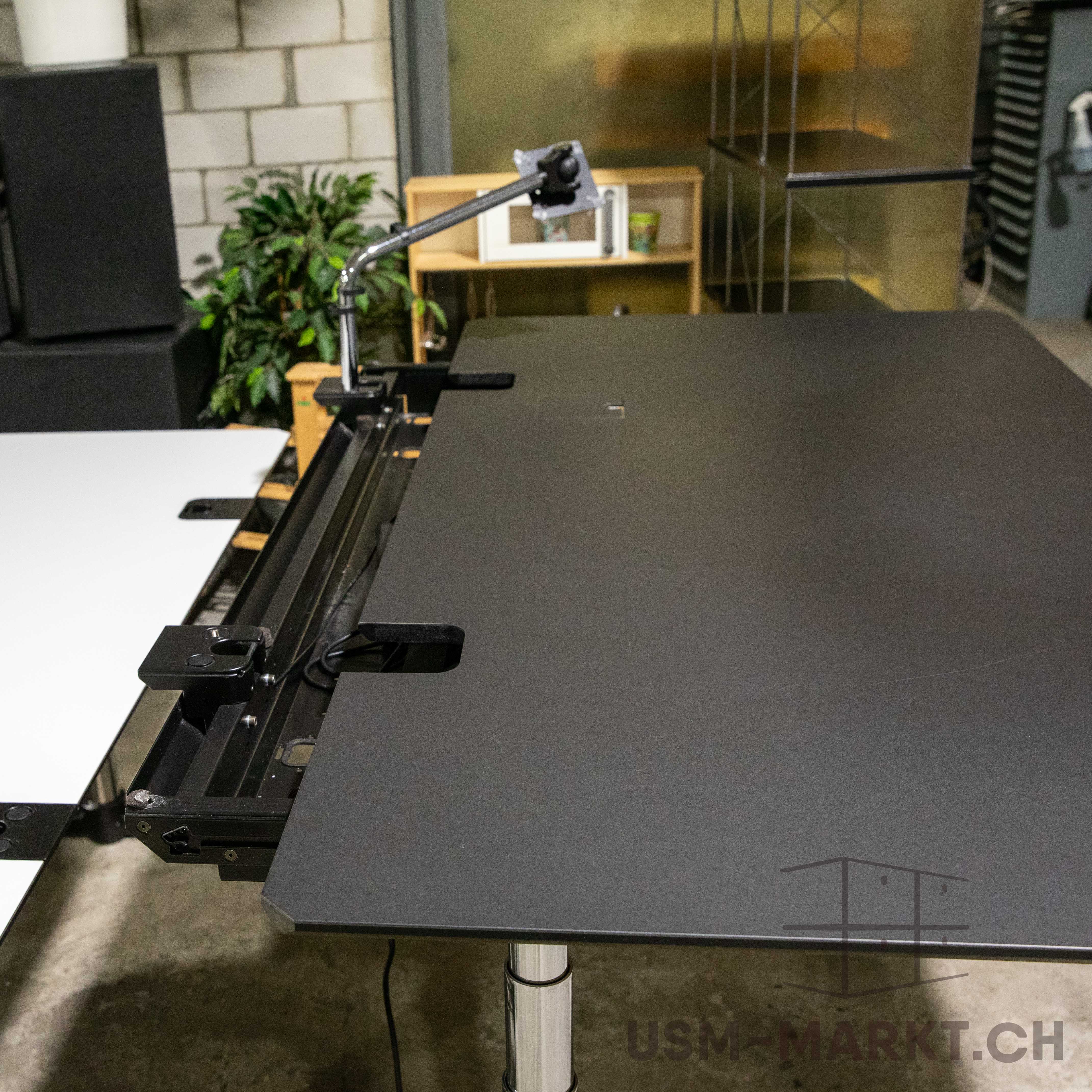 Elektrisch höhenverstellbarer USM Kitos E Plus Schreibtisch mit mattschwarzer Linoleumplatte und leichten Gebrauchsspuren. Integriertes Kabelmanagement mit Monitorarmhalterung an der Hinterkante, inkl. Kabelklappe rechts hinten.