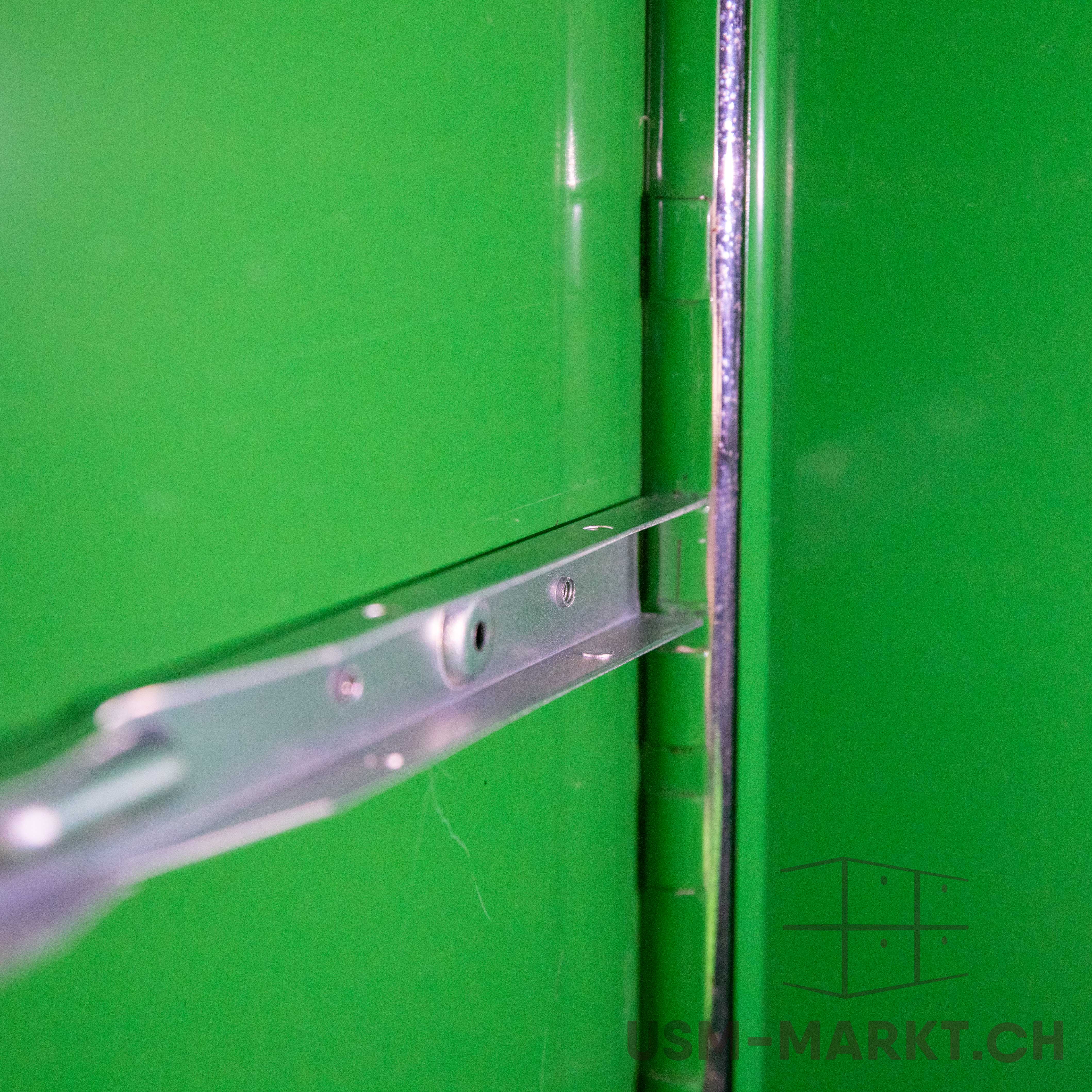 Close-up of a shiny silver metal strut, fixed diagonally to a bright green surface. Secured with a round screw or rivet, it reflects light. To the right, a vertical metal edge of a sturdy structure.