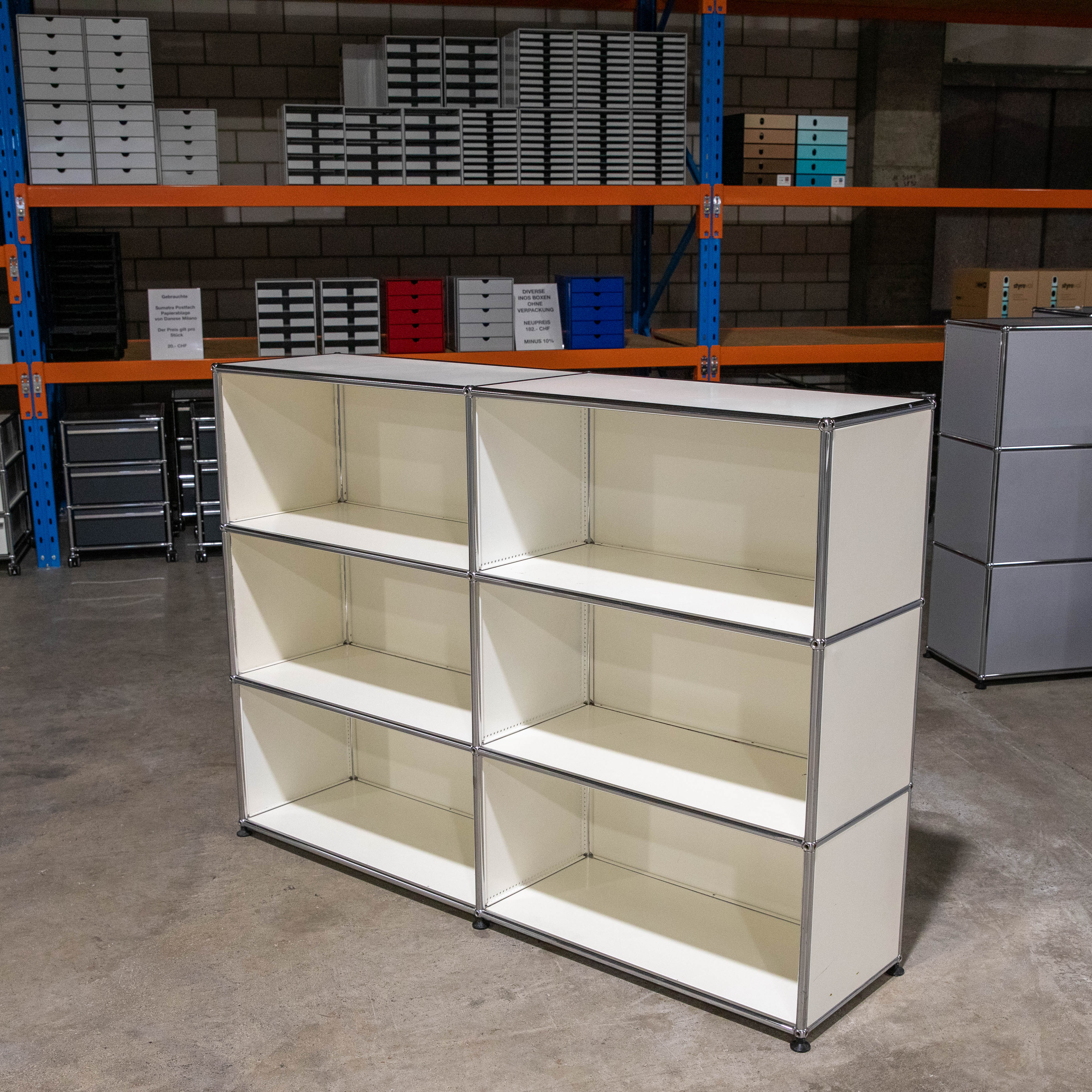Leeres, weißes modulares Regal mit sechs offenen Fächern auf grauem Betonboden. Im Hintergrund hohe blaue und orange Lagerregale mit vielen kleinen grau-schwarzen und bunten Bürospeicherschubladen. Ziegelwand sichtbar.