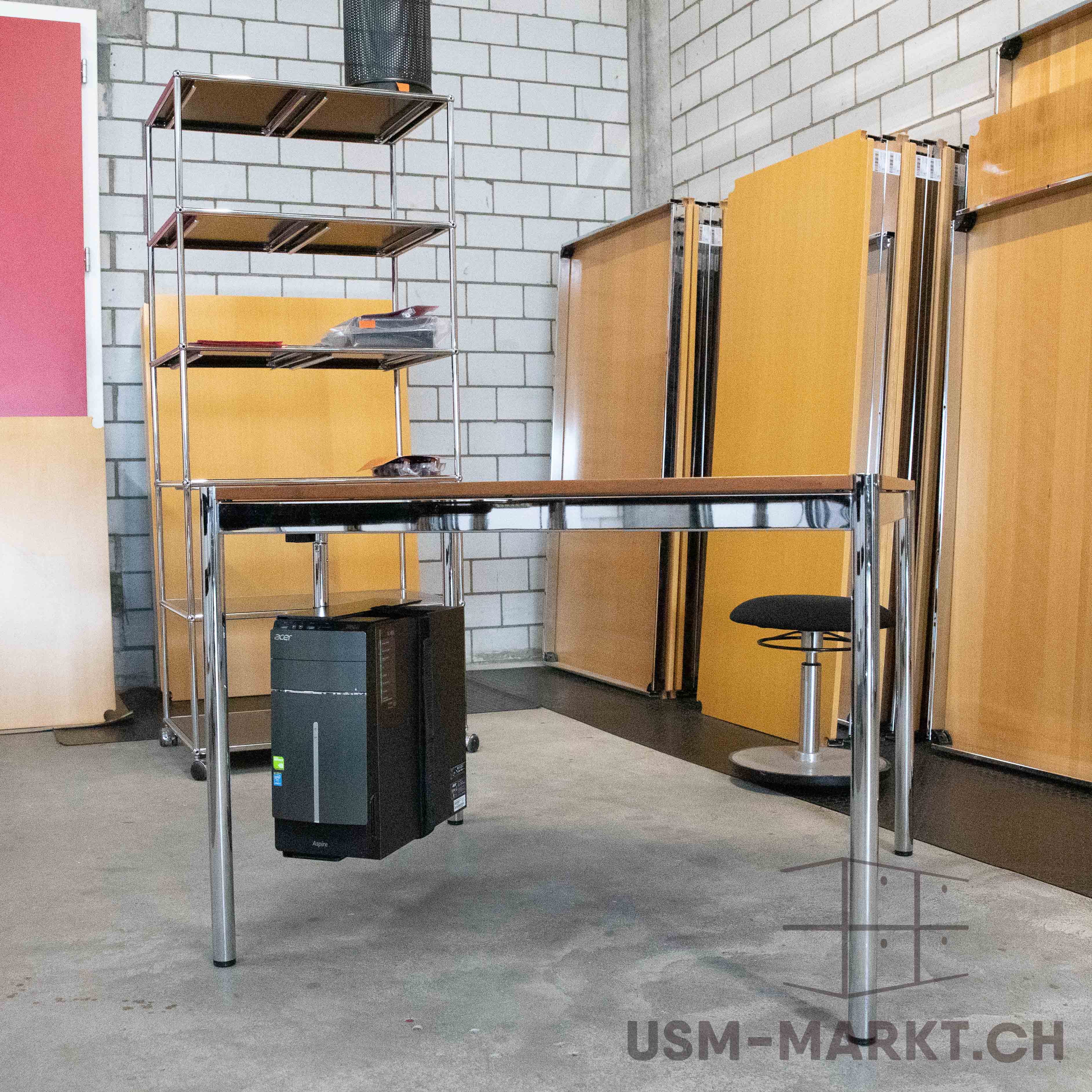 Modern office desk with dark wooden top and chrome legs in a storeroom. Left: chrome shelving unit with four dark shelves holding papers and a wastepaper bin. Under the desk is a black computer. Rear: wooden panelling and brick wall.