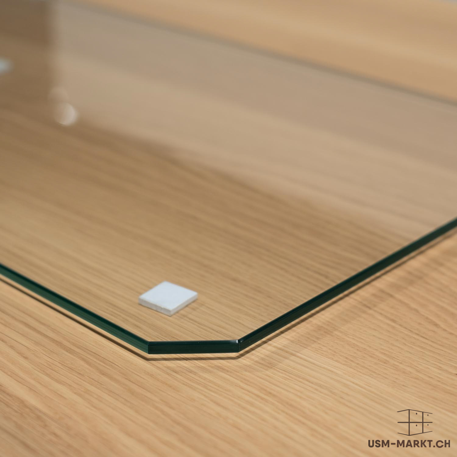 Piastra in vetro trasparente con bordo dai riflessi verdastri su una superficie in legno marrone chiaro venato. Un angolo leggermente arrotondato poggia su un distanziatore quadrato bianco, sollevando la lastra. La luce soffusa si riflette.