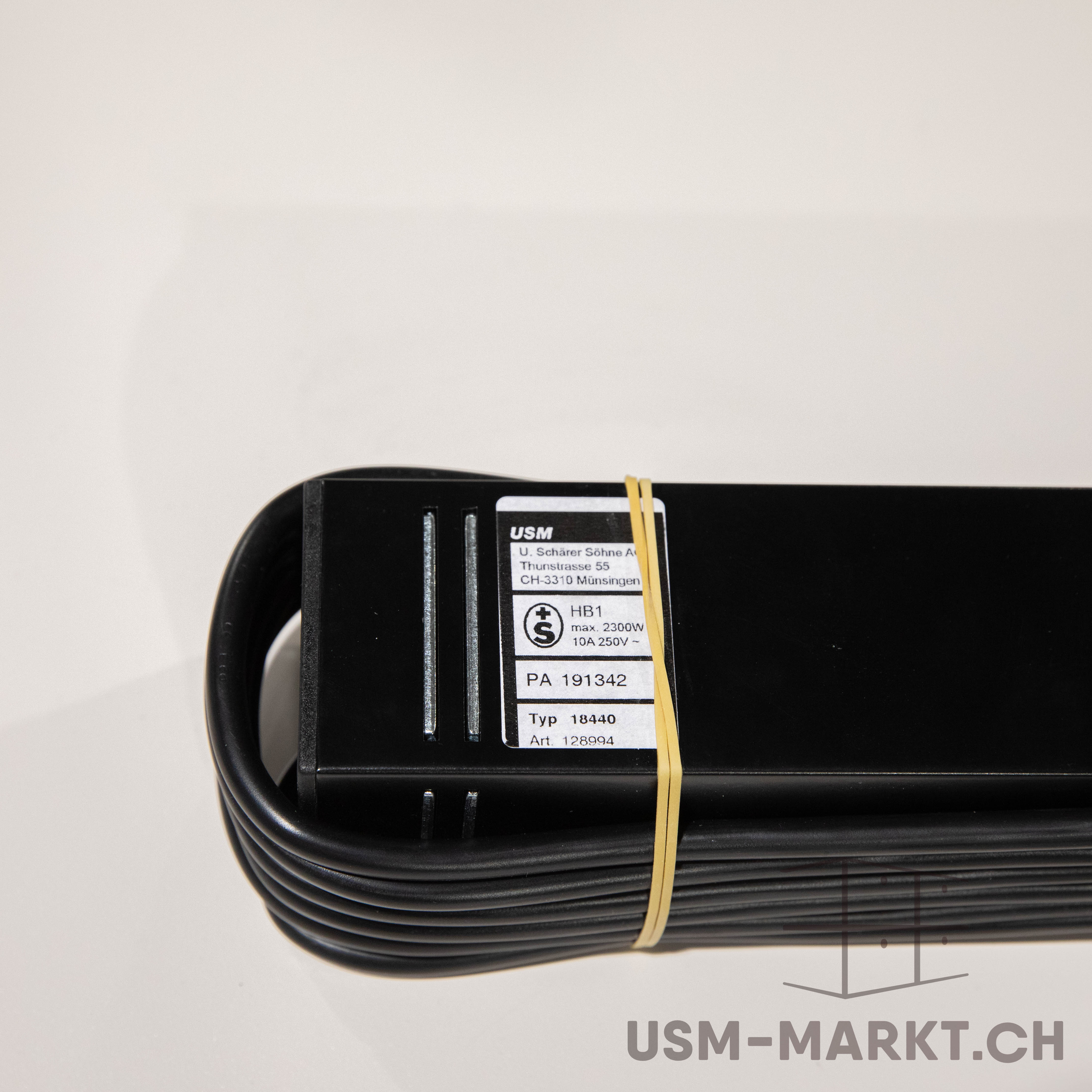 A black, unused USM power strip with a neatly coiled power cable held in place by a yellow rubber band. A white label shows manufacturer details and technical specifications such as 2300W, 10A, 250V and item numbers.