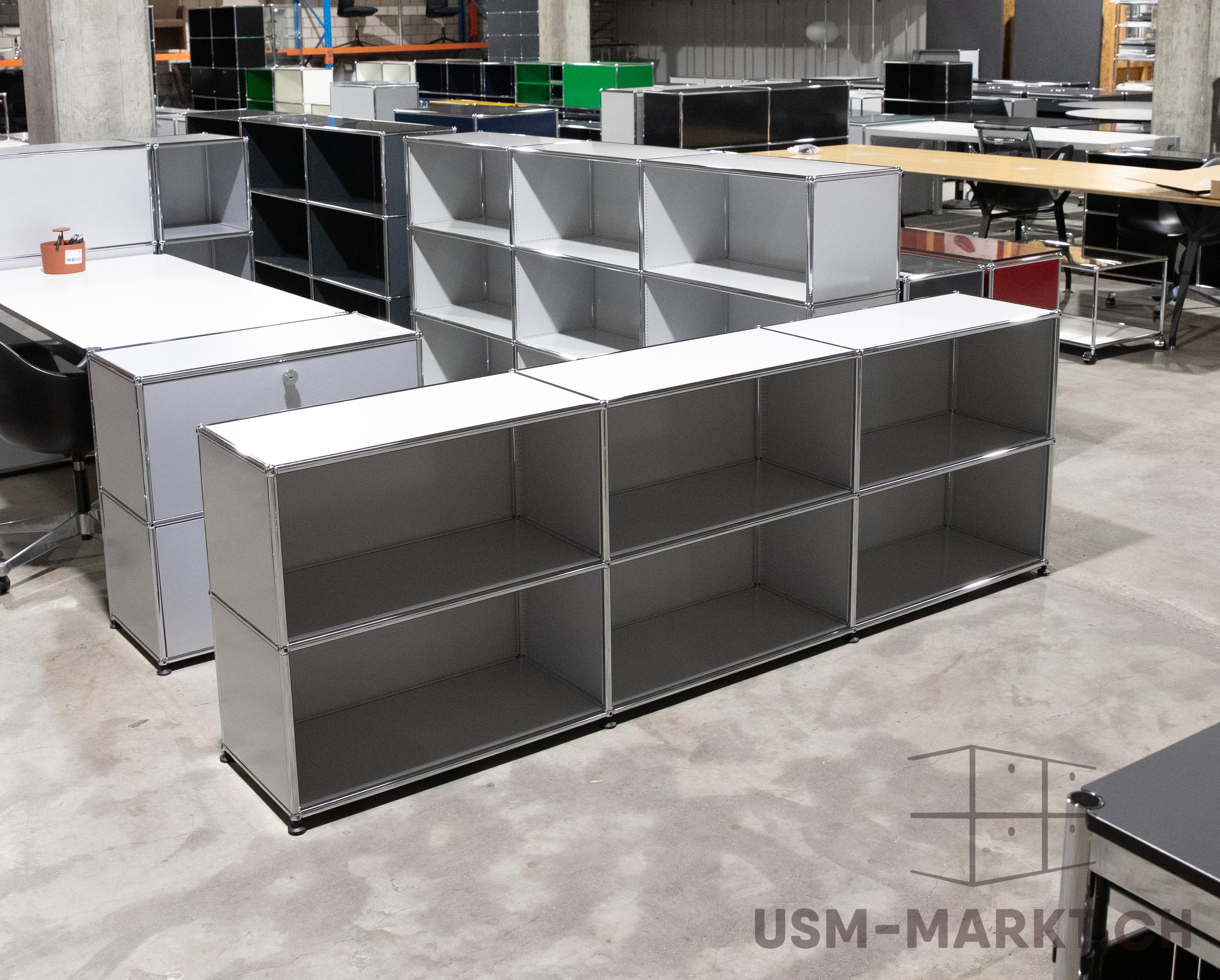 Weitläufige Halle mit modularen USM Haller Möbeln in Grau, Schwarz, Weiß, Rot und Grün. Vorn offene Regale und Sideboards, teils als Bürotische mit Stühlen. Betonboden und hohe Decken: industrieller Showroom/Lager.
