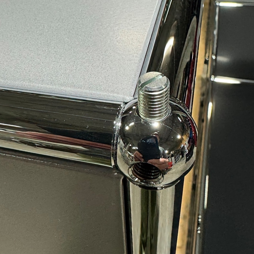 A glossy chrome corner unit with a light grey textured top, edged with silver. A spherical chrome joint connects parts, secured with a slotted screw. Reflective, showing a distorted reflection of a person or surroundings. Side is dark grey.