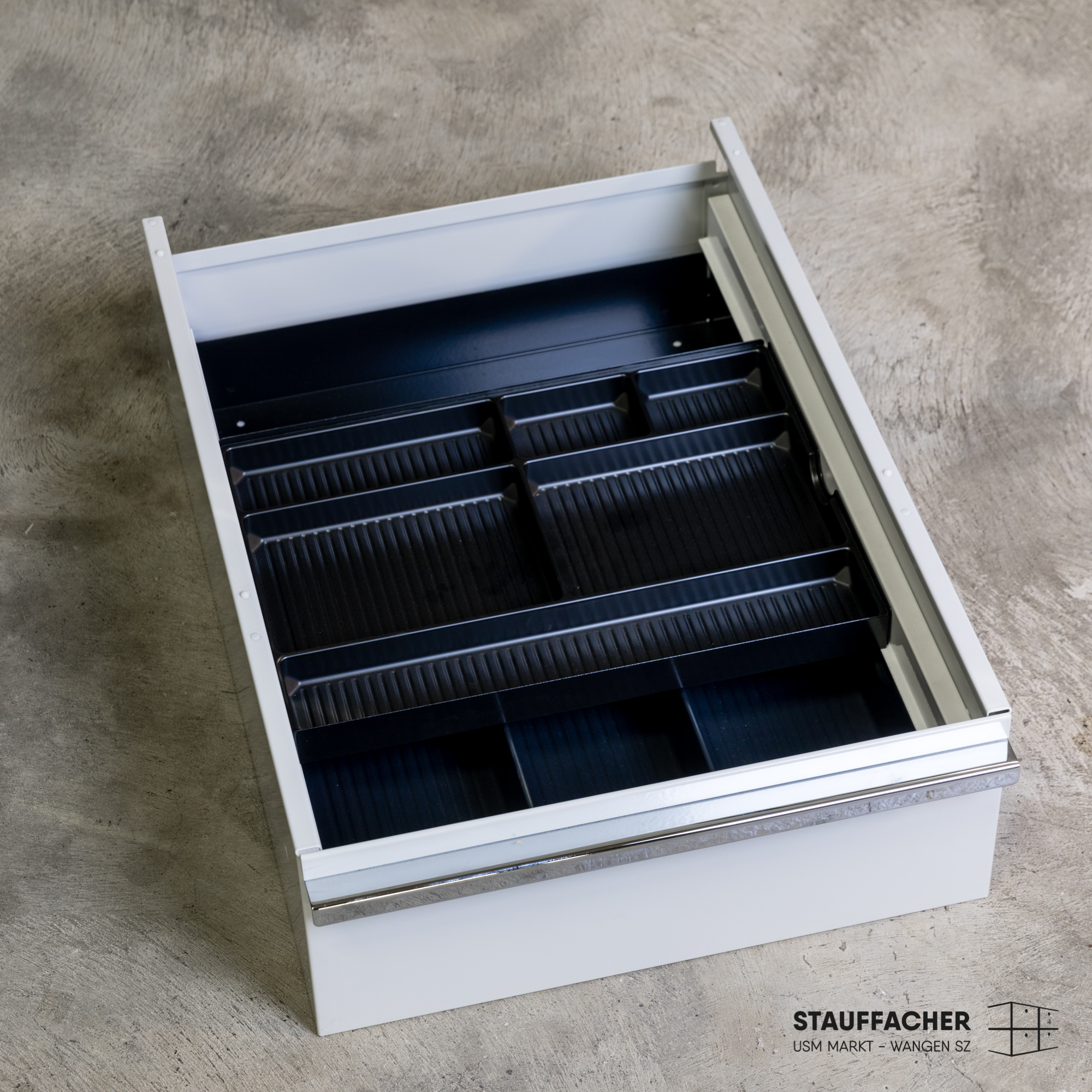 Cassetto bianco con portaposate a coste blu scuro dotato di diversi scomparti per organizzare gli utensili. Con maniglia argentata sul davanti, poggia su una base in cemento grigio, leggermente aperto.