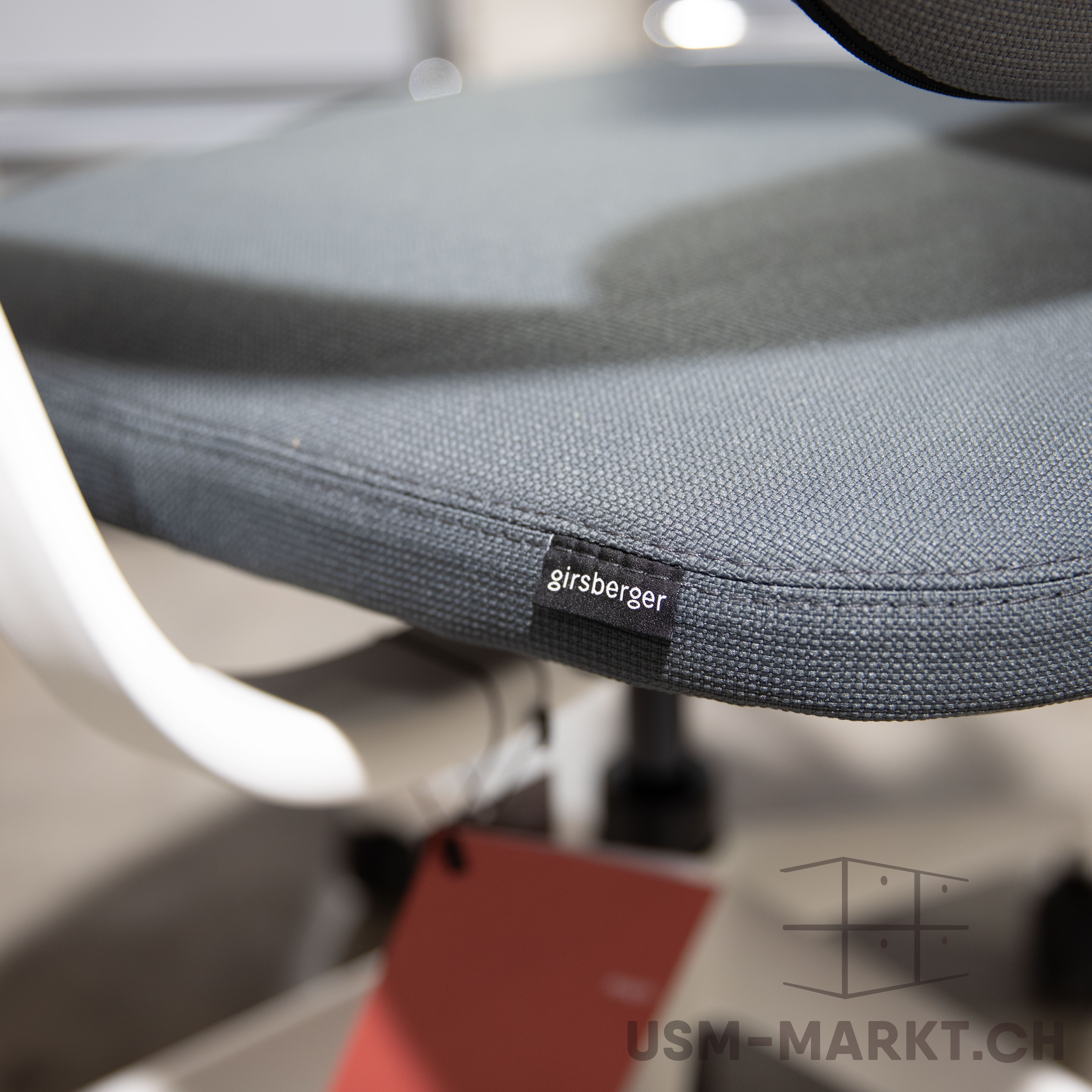 Gros plan sur l’assise d’une chaise de bureau grise à texture tissée fine. Sur le bord avant du coussin, une étiquette noire « Girsberger » en lettres blanches. À gauche, partie d’accoudoir blanc avec étiquette rouge.
