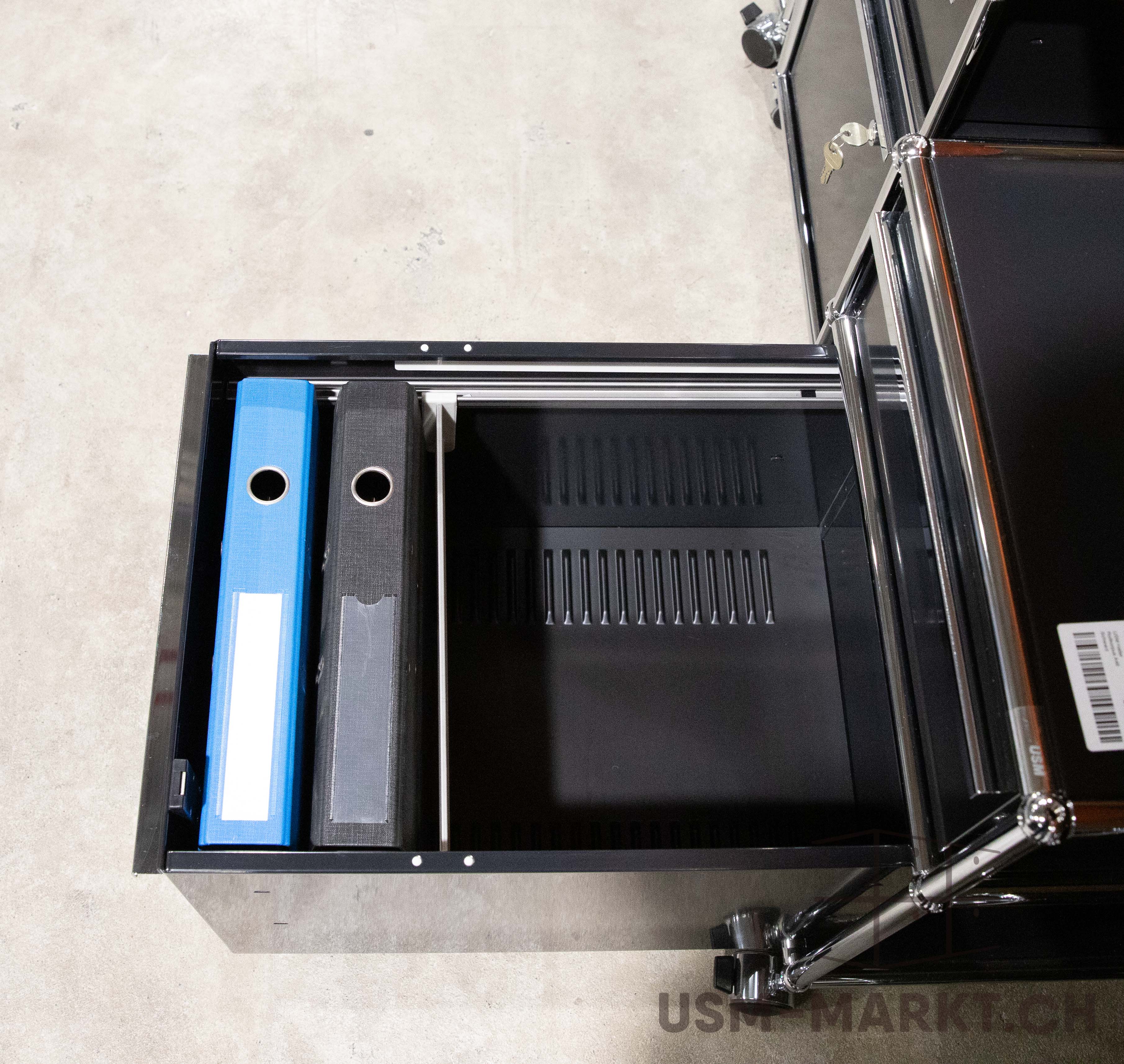 Offene Schublade eines schwarzen Rollcontainers mit blauem und grauem Aktenordner. Die rechte Hälfte ist leer und zeigt die schwarze Innenverkleidung mit horizontalen Schlitzen. Oben rechts sind Rahmen und Schlüssel sichtbar.