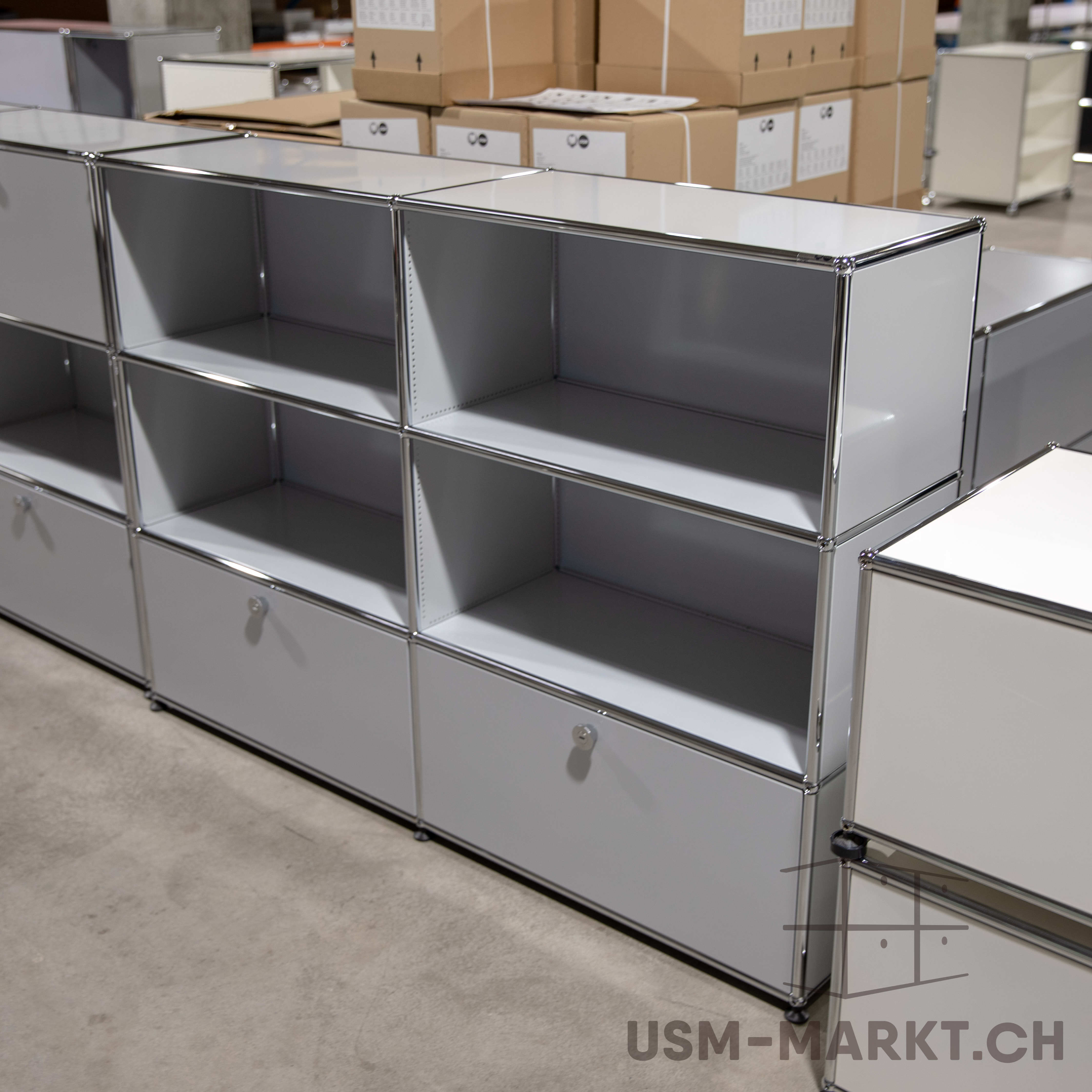 Lungo sistema di scaffalature modulari composto da elementi grigio chiaro con telai cromati su pavimento in cemento chiaro. Scomparti aperti e cassetti chiusi con maniglie metalliche rotonde. Sullo sfondo, scatole impilate e altre scaffalature.