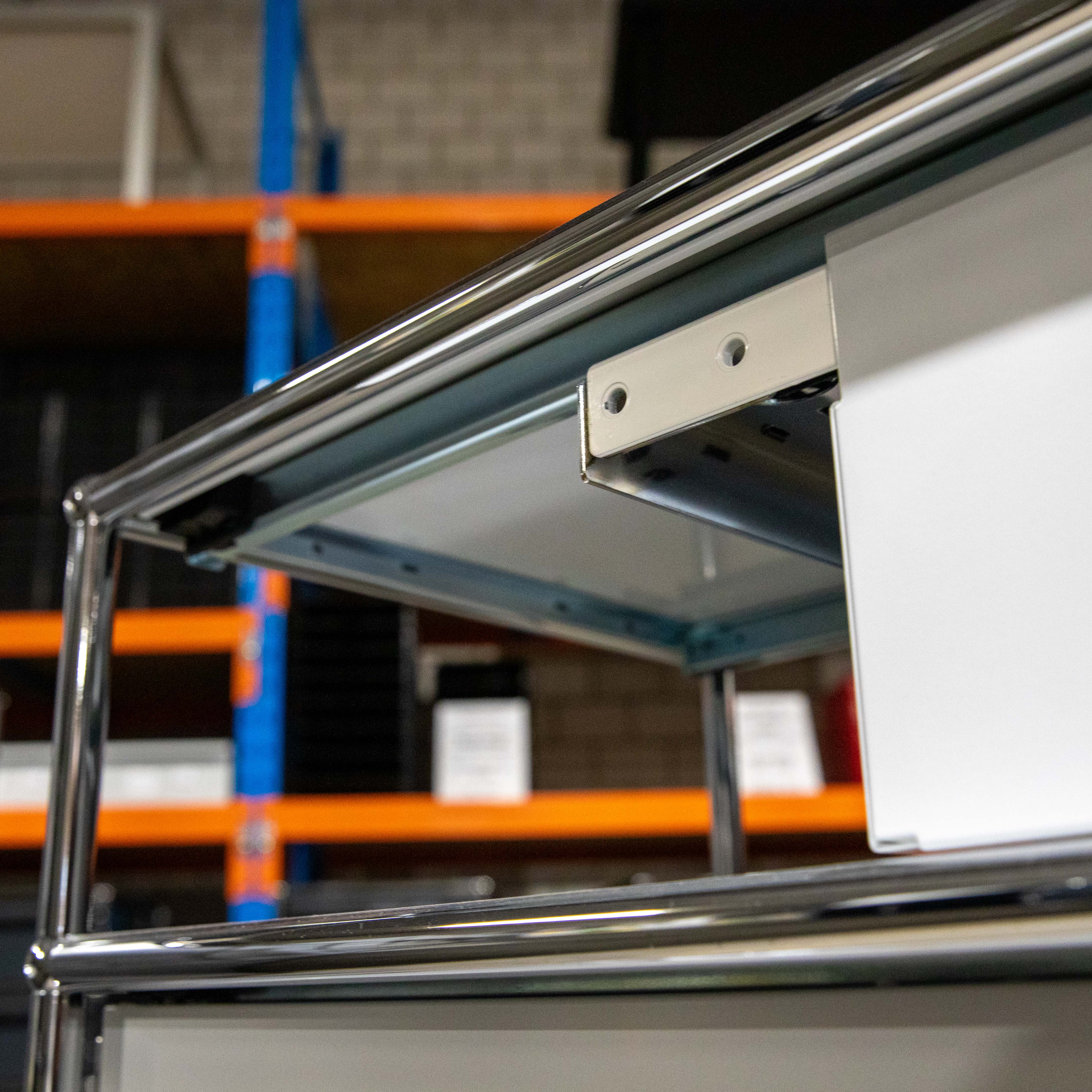 Primo piano di una struttura con telaio cromato lucido che sostiene un pannello bianco. Sulle superfici metalliche sono visibili dei riflessi. Sullo sfondo sfocato si intravedono scaffali di magazzino di colore arancione e blu.