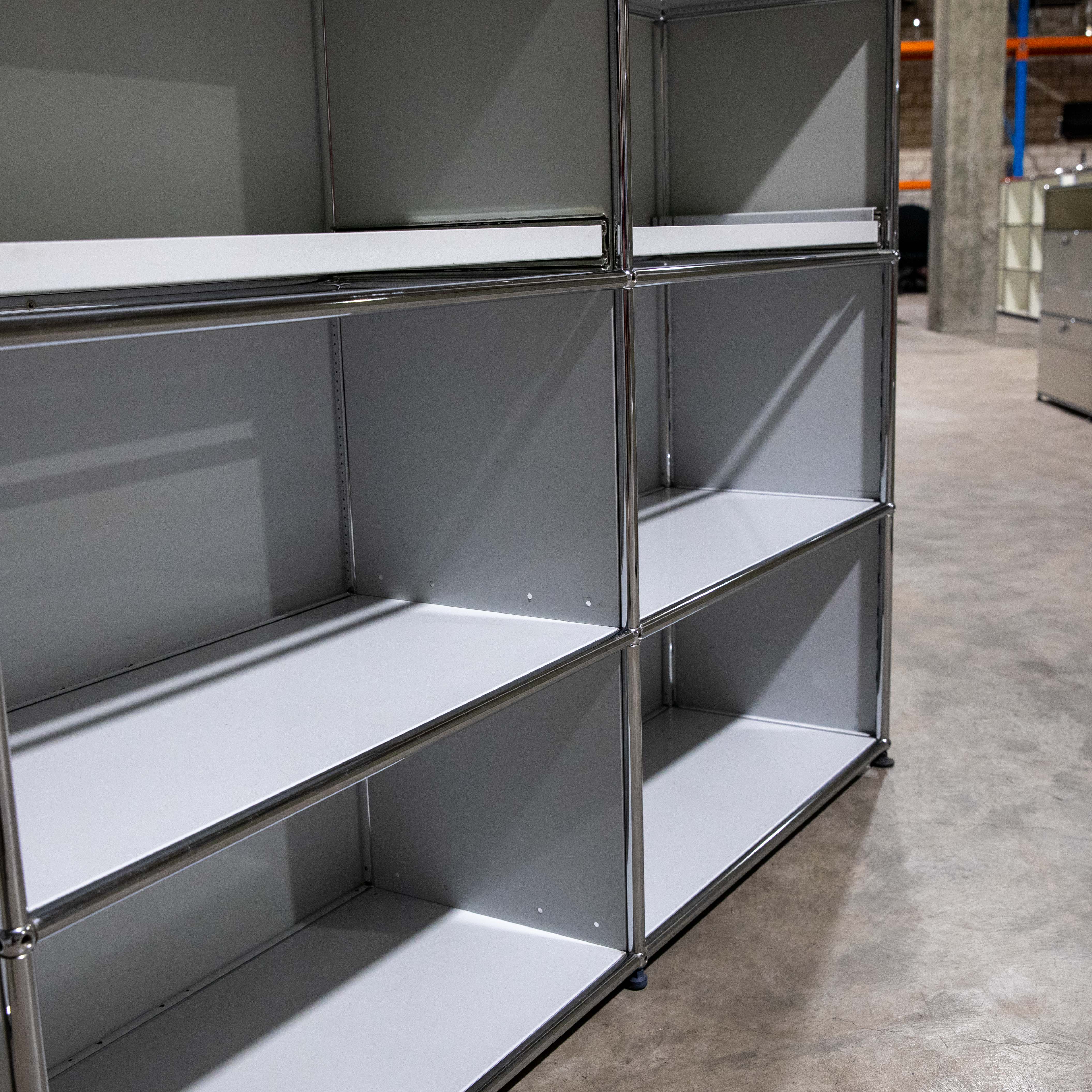 Second-hand USM Haller shelving unit in light grey, modular, with two columns and three rows of open compartments, framed by polished chrome steel tubes. The pull-out shelf at the top left is slightly open. Standing on a grey concrete floor.