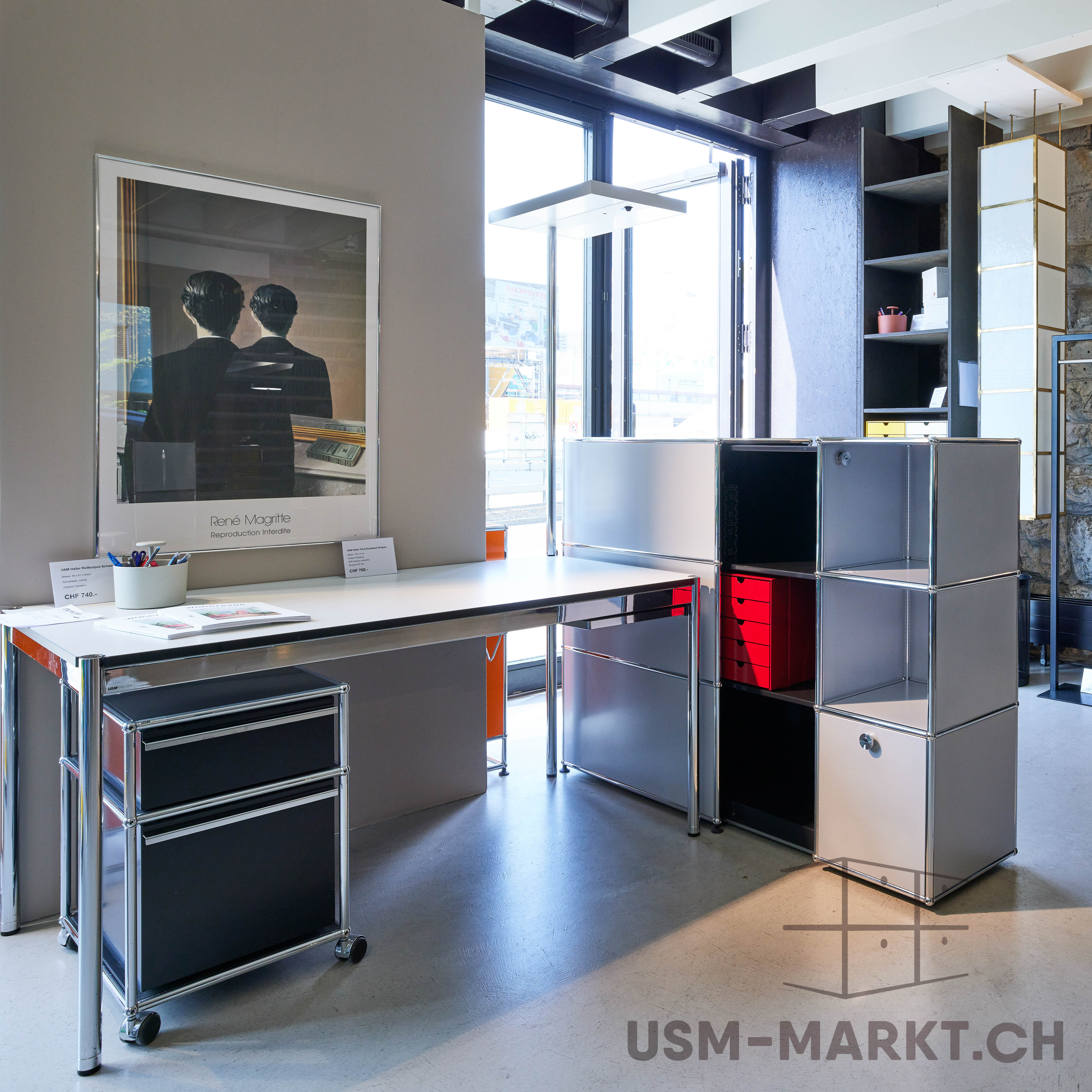 Modernes Büro mit weissem USM Haller Schreibtisch und schwarzem Rollcontainer. An der Wand ein Magritte-Poster: Mann von hinten vor Spiegel, der ebenfalls den Rücken zeigt. Rechts silbernes USM Regal mit roter Schublade.