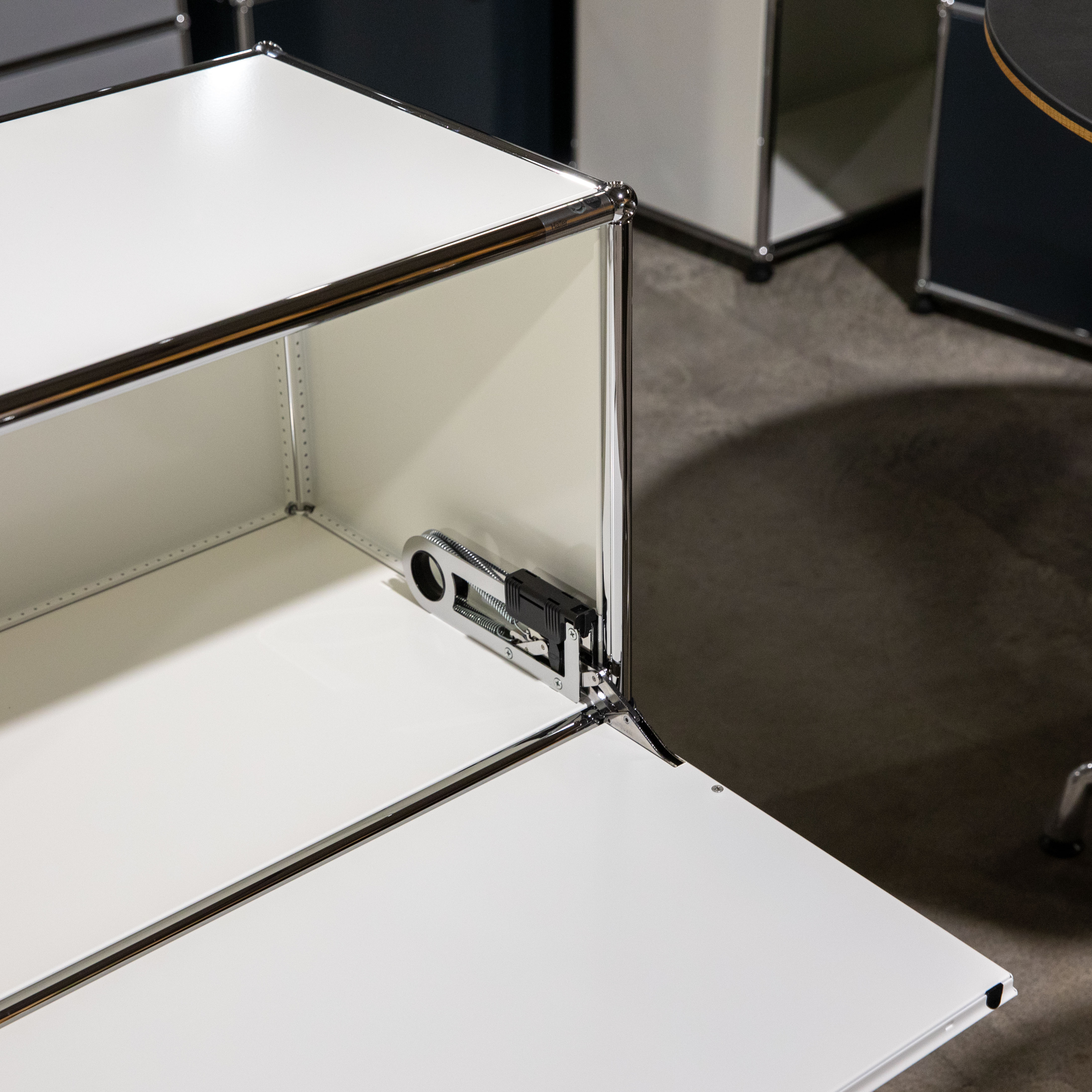 Nahaufnahme eines offenen Fachs eines weißen USM Haller Sideboards. Die weiße Klapptür ist nach unten geöffnet und zeigt den silbernen Schließmechanismus mit Federn und schwarzem SilentClose-Dämpfer. Innen sind perforierte Rückwände sichtbar.