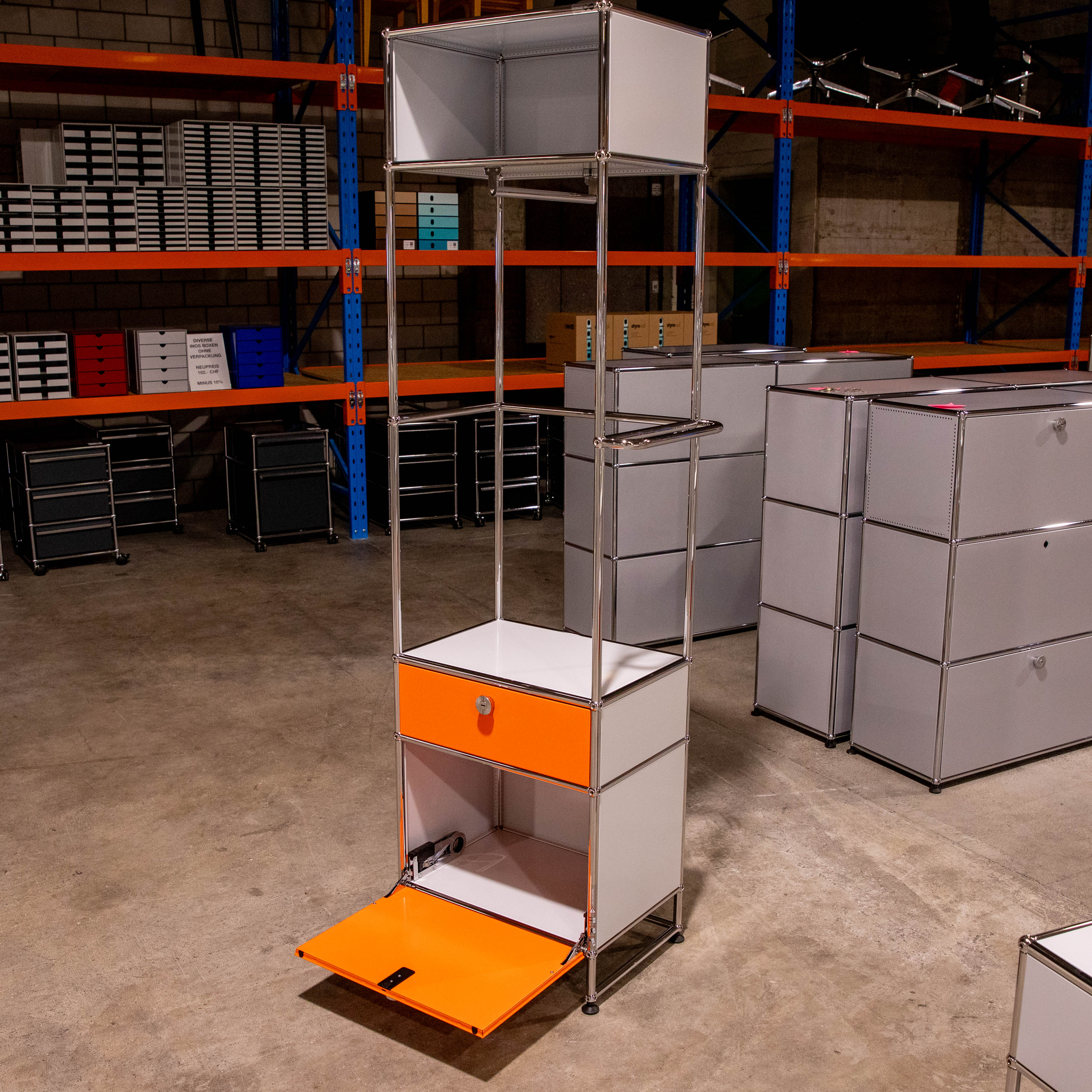 Gebrauchte USM Haller Garderobe in Lichtgrau/Orange: hohes modulares Regal mit Chromstangen. Orange Schublade, darunter offene Klapptür. Oben offenes Fach, rechts Kleiderstange. Auf Betonboden in Lagerhalle.