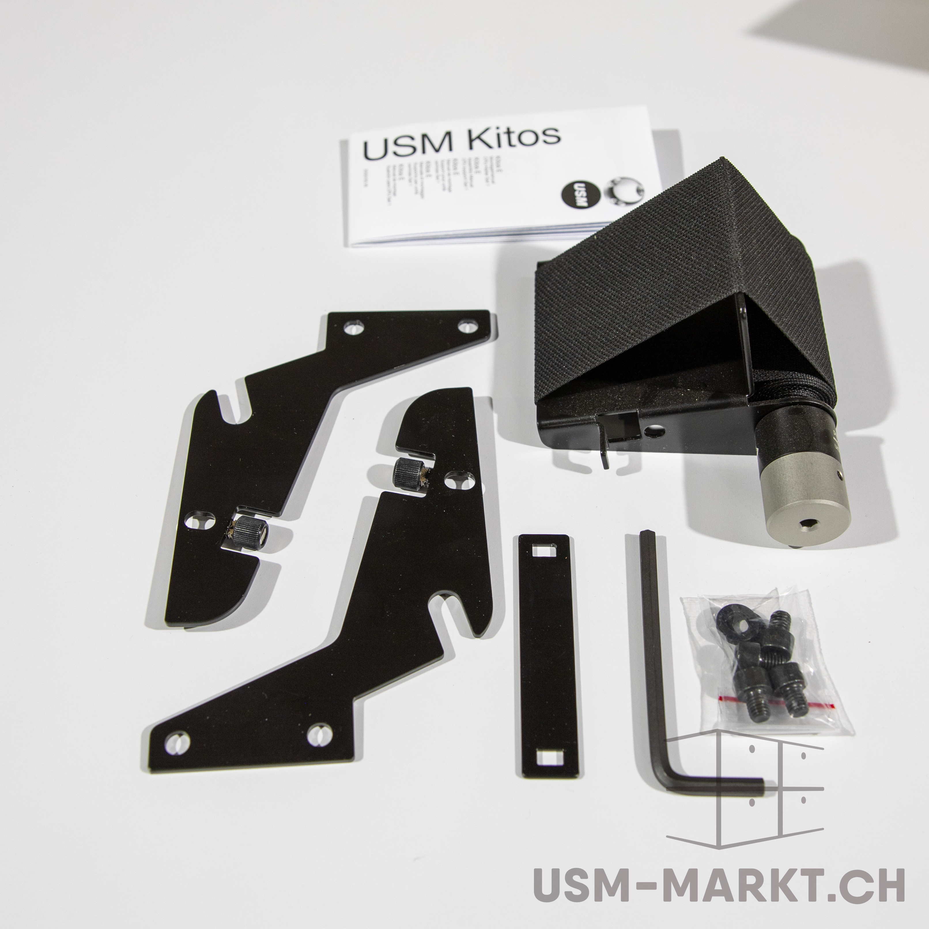 USM Kitos-Komponenten auf weißem Hintergrund: gefaltetes Handbuch, mehrere schwarze Metallhalterungen, schwarze Stoffabdeckung mit zylindrischem Anschluss, Innensechskantschlüssel sowie Tüte mit Schrauben/Unterlegscheiben.