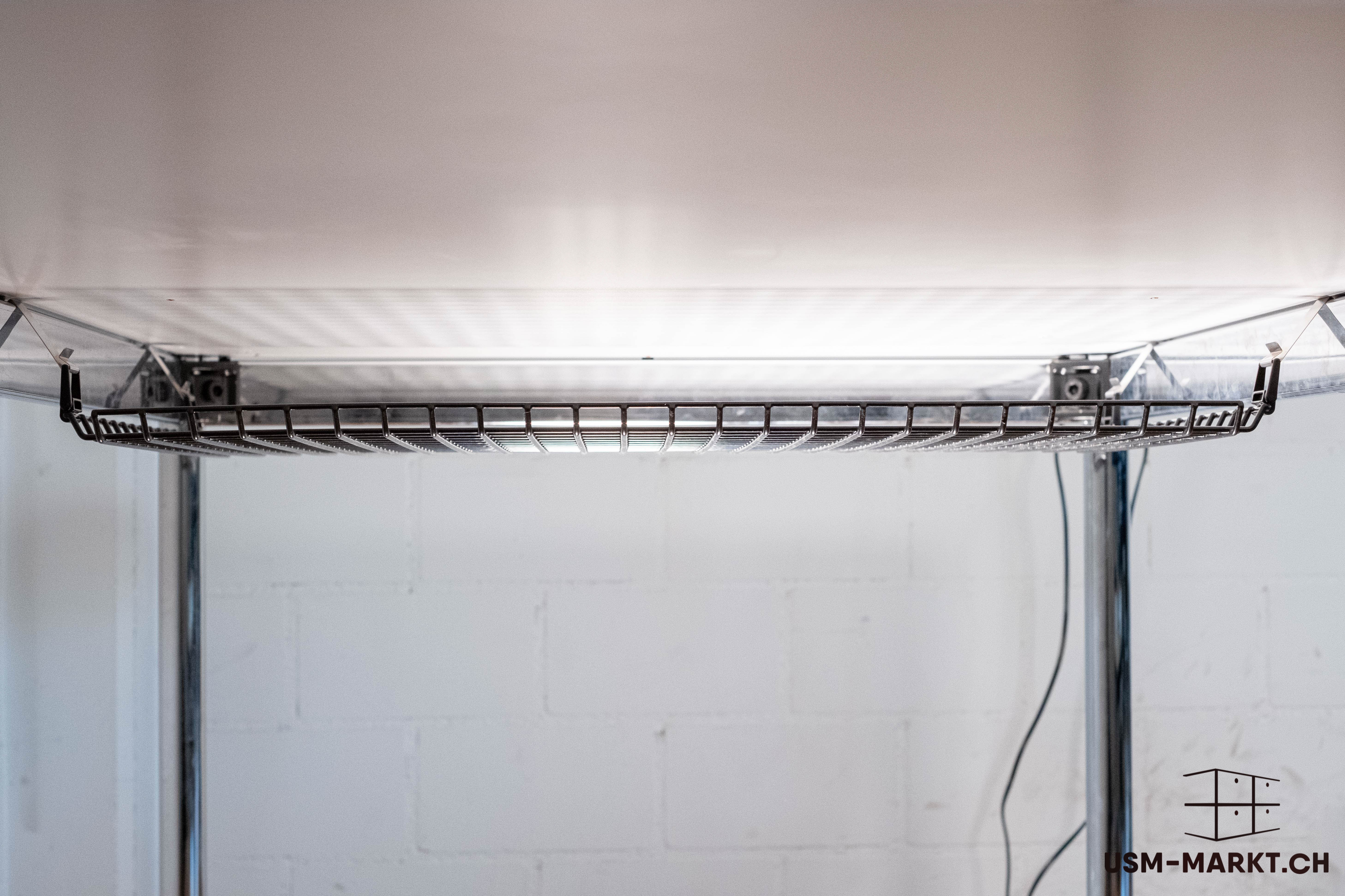 Schwarze, gitterförmige Kabelwanne unter heller Tischplatte zur Kabelorganisation, mit mehreren Halterungen befestigt. Im Hintergrund weisse Ziegelwand und glänzende Metalltischbeine. Rechts hängt ein dunkles Kabel herab.