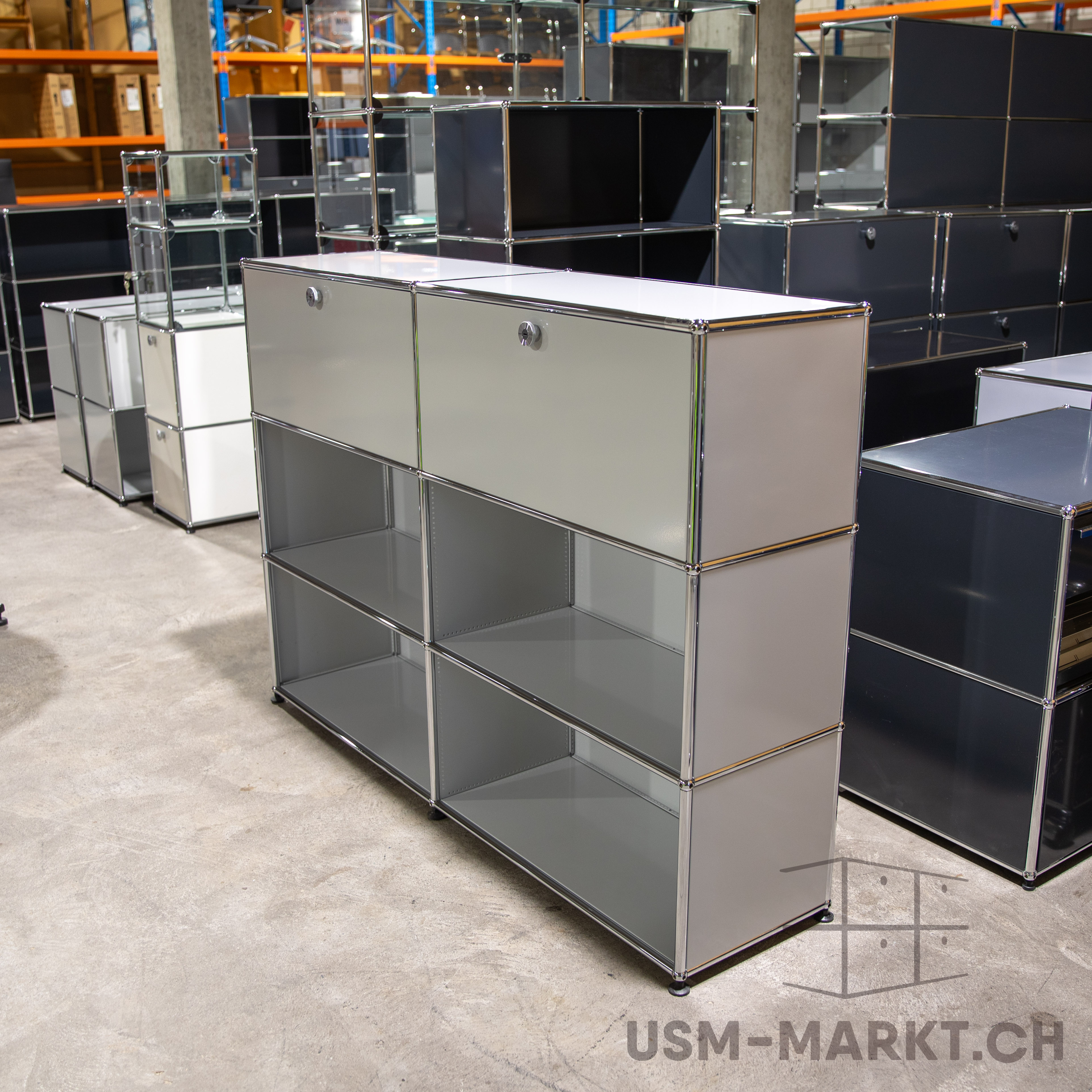 Modulare USM Haller Möbel in Lager oder Showroom. Vorn zwei hellgraue Sideboards mit offenen Regalfächern und Türen. Hinten weitere Elemente in Grautönen mit Glaseinsätzen auf Betonboden.