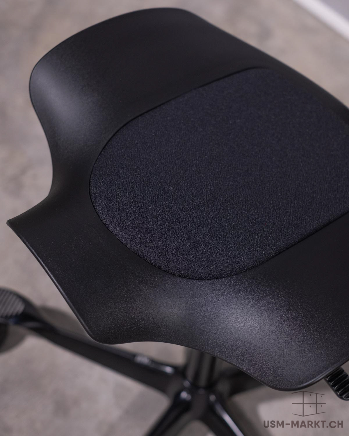 Primo piano della seduta nera dal design ergonomico di una moderna sedia da ufficio. Rivestimento in tessuto scuro testurizzato al centro, circondato da plastica liscia e opaca. La sedia poggia su una superficie scura ed è dotata di rotelle.