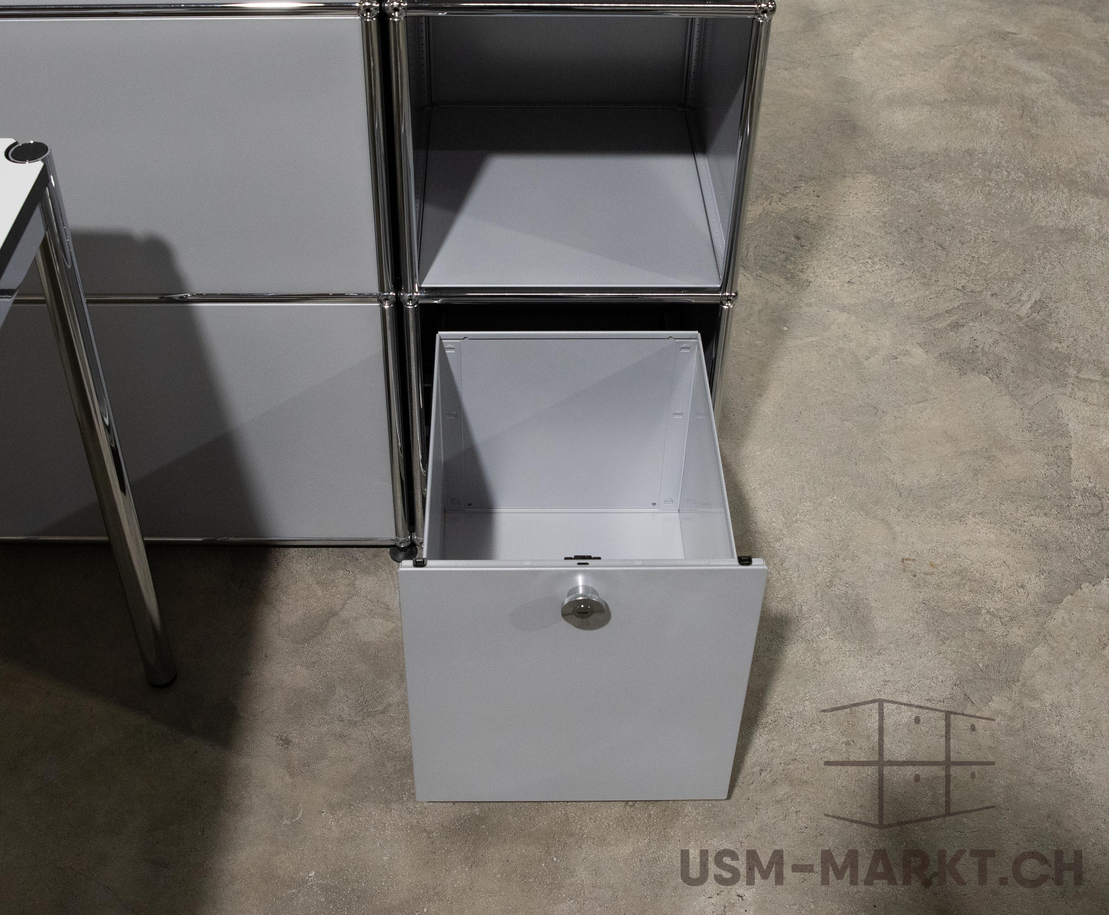 Hellgraues USM Haller Sideboard mit modularen Fächern. Oben rechts ein offenes Fach, darunter eine ganz ausgezogene Schublade mit silbernem Schloss. Verbunden mit glänzenden Chromrohren, auf grauem Betonboden.