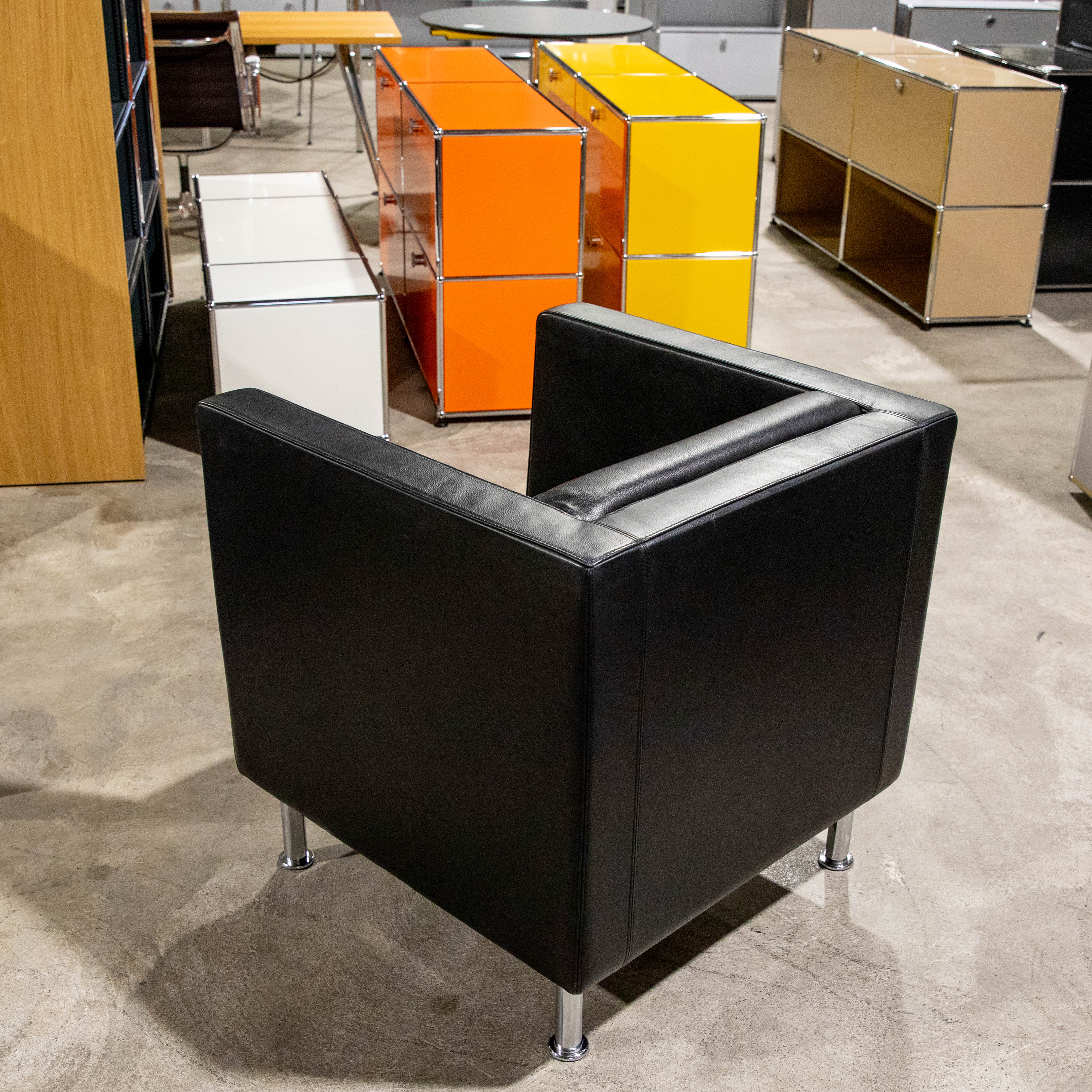 A stylish black leather armchair with shiny chrome legs stands at the front on a light grey concrete floor. In the background are modern modular furniture, eye-catching orange and yellow cupboards, and beige and white shelving units.