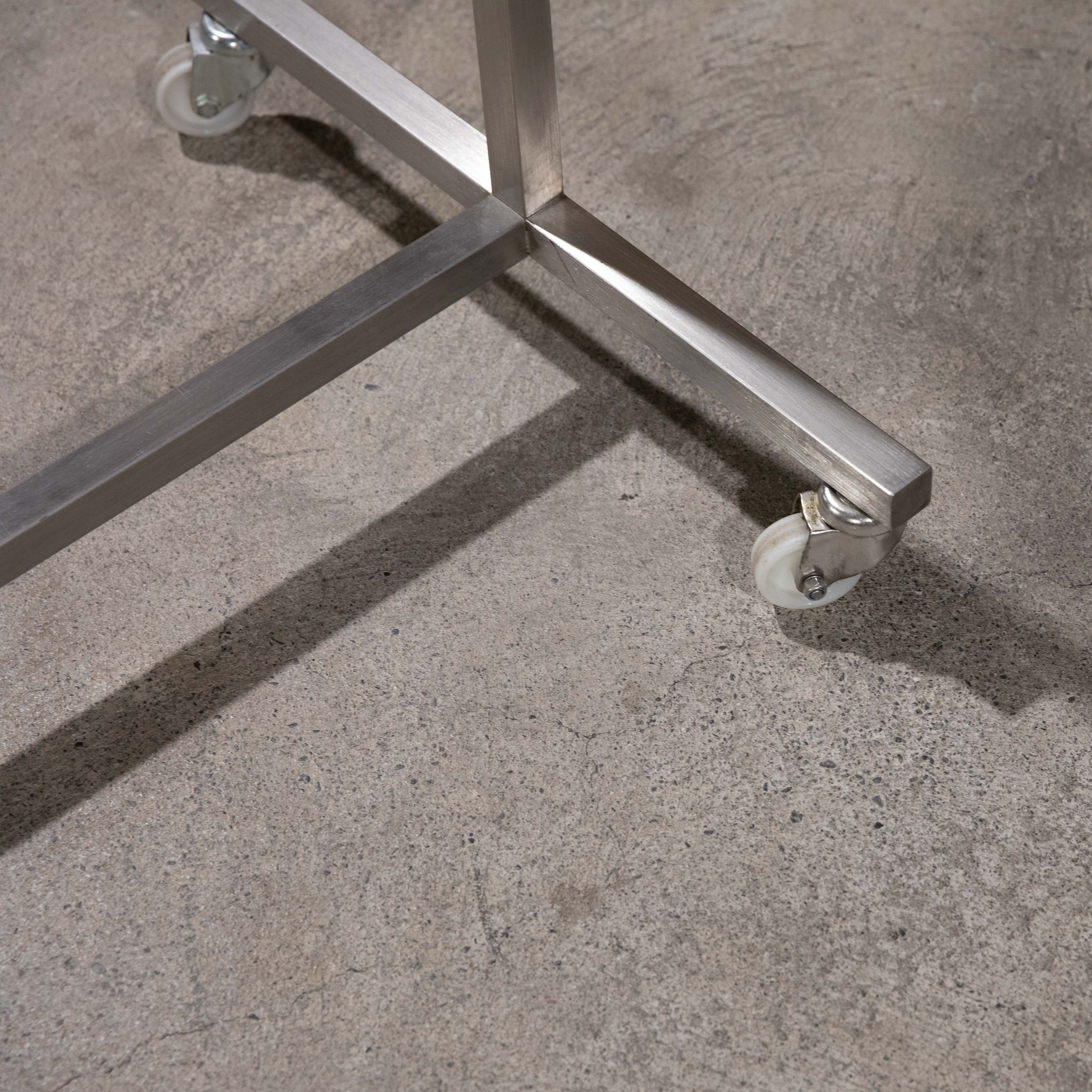 Detailansicht des massiven Chromstahl-Gestells eines rollbaren Kleiderständers. Stabile, viereckige Metallstreben bilden die Basis, mit weißen, leicht transparenten Lenkrollen an den Enden. Steht auf hellgrauem Betonboden; Schatten sichtbar.