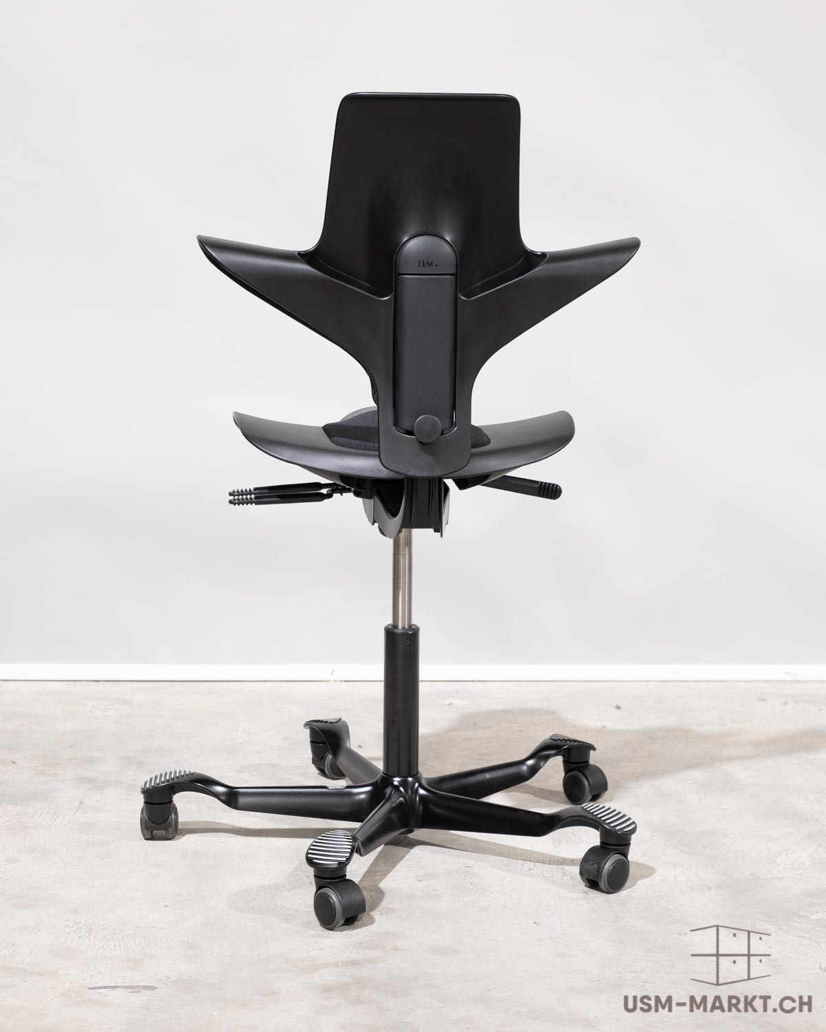 Sedia da ufficio nera HÅG moderna, vista da dietro, con schienale sagomato e seduta a forma di ali. Base a stella a cinque bracci con rotelle e poggiapiedi a strisce. Su cemento grigio chiaro davanti a parete bianca.