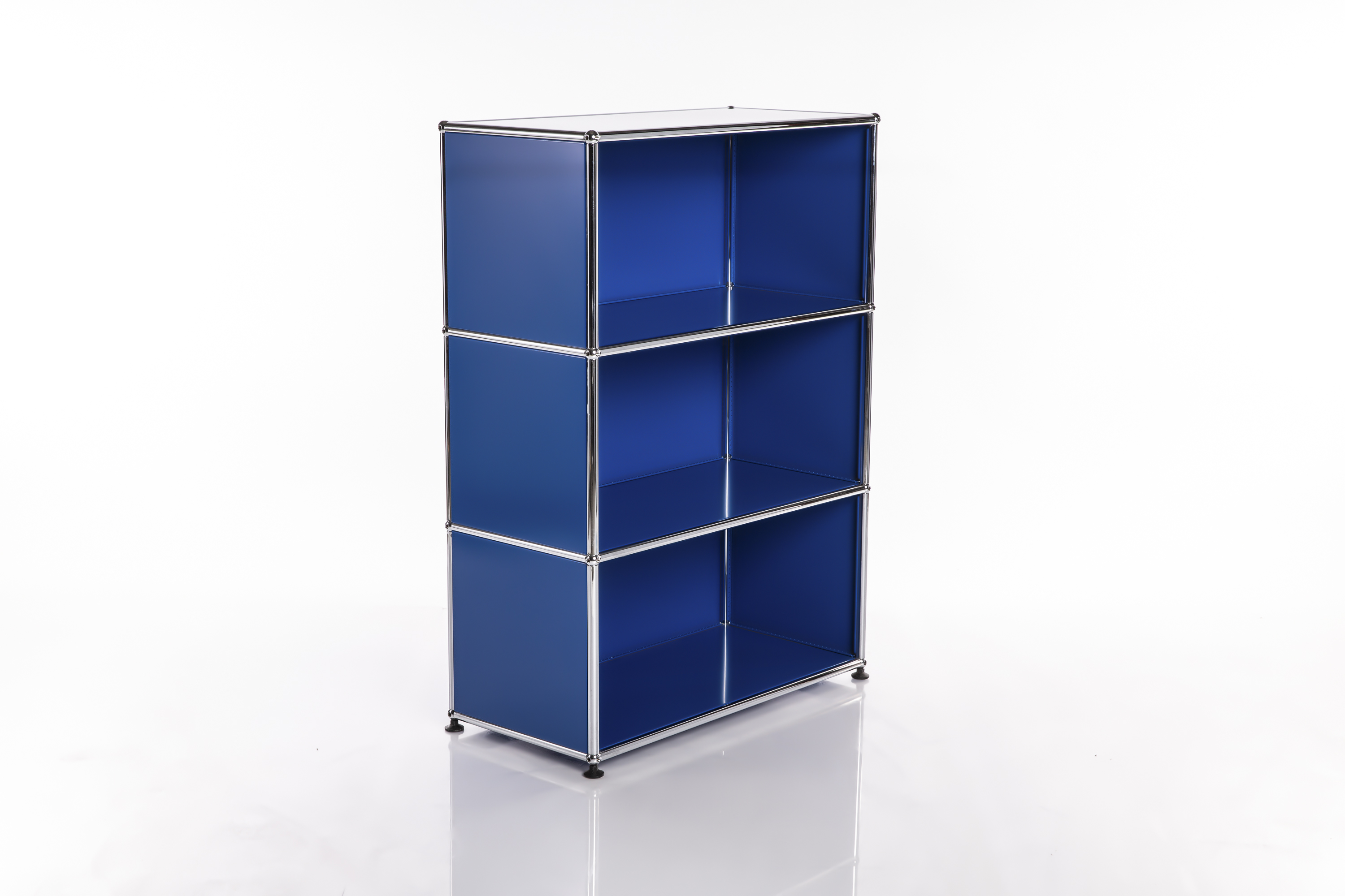 Scaffale minimalista e modulare in un brillante blu reale con tre ripiani aperti sovrapposti. Struttura in cromo lucido con angoli arrotondati. Poggiato su piccoli piedini scuri, proietta una delicata ombra sul fondo bianco lucido.
