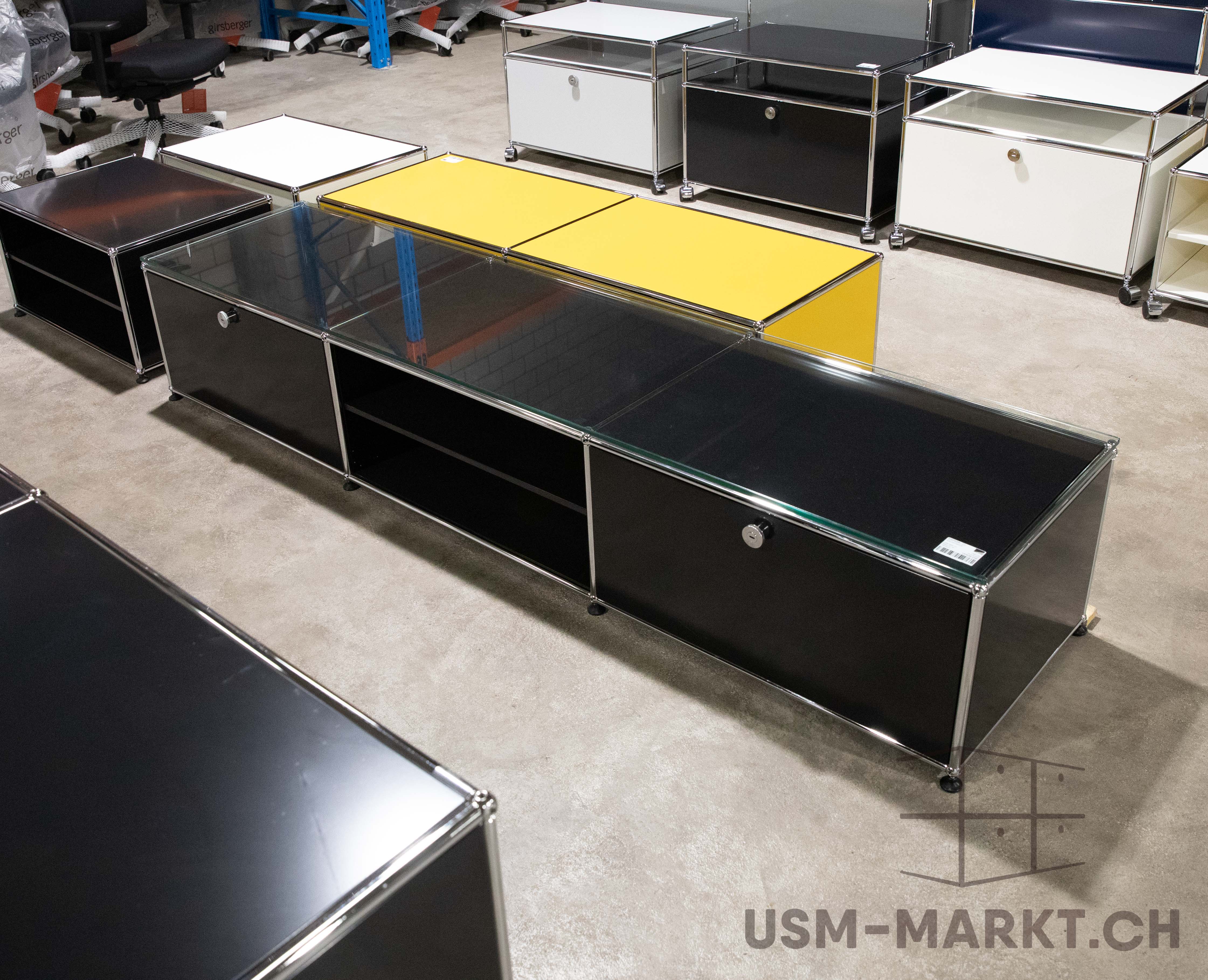 A long, black USM Haller sideboard with a glass top and chrome tubes on a concrete floor. Two open compartments and two lockable drawers. In the background, modular furniture in black, white and yellow, as well as boxed office chairs.